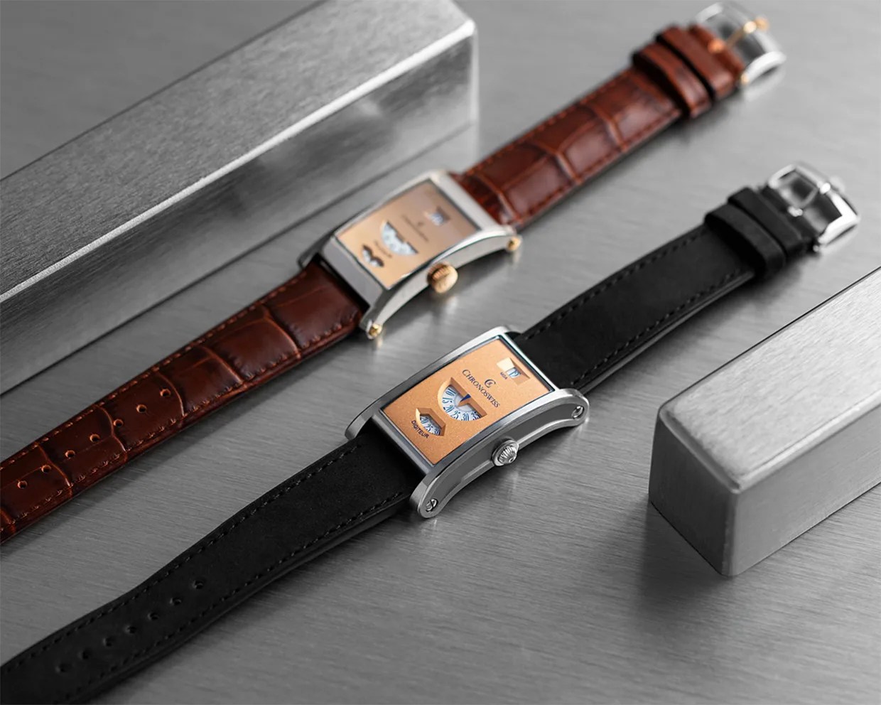 Two rectangular wristwatches with orange dials, one with a black leather strap and the other with a brown crocodile leather strap, on a metallic surface.