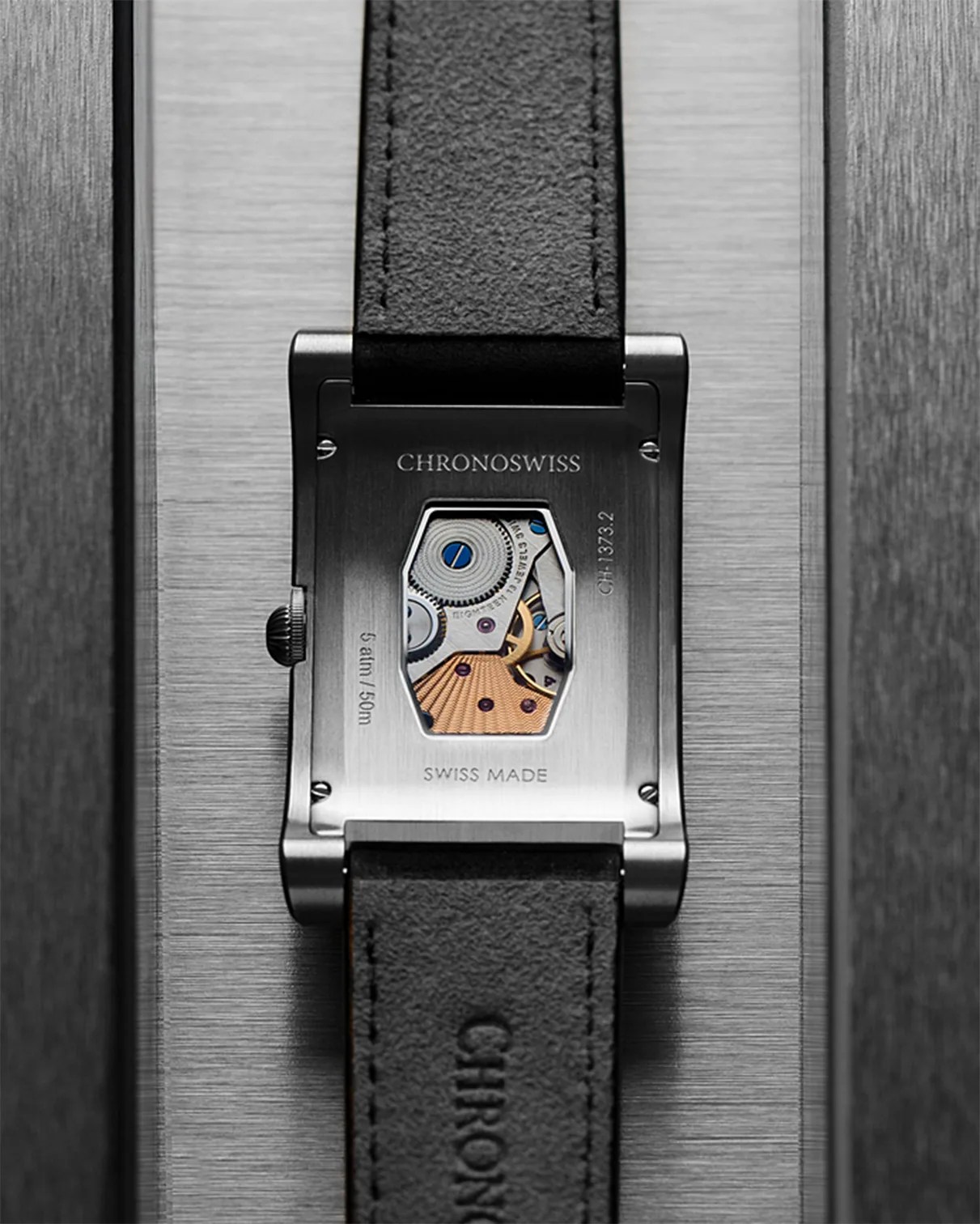 Back of a rectangular Chronoswiss watch with visible mechanical movement and black leather strap on a brushed metal surface.