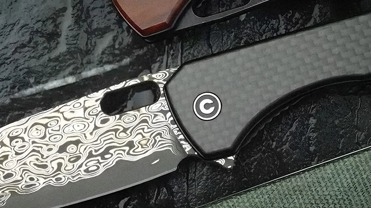 Close-up of a folding knife with a patterned Damascus steel blade and a carbon fiber handle featuring a circular logo.