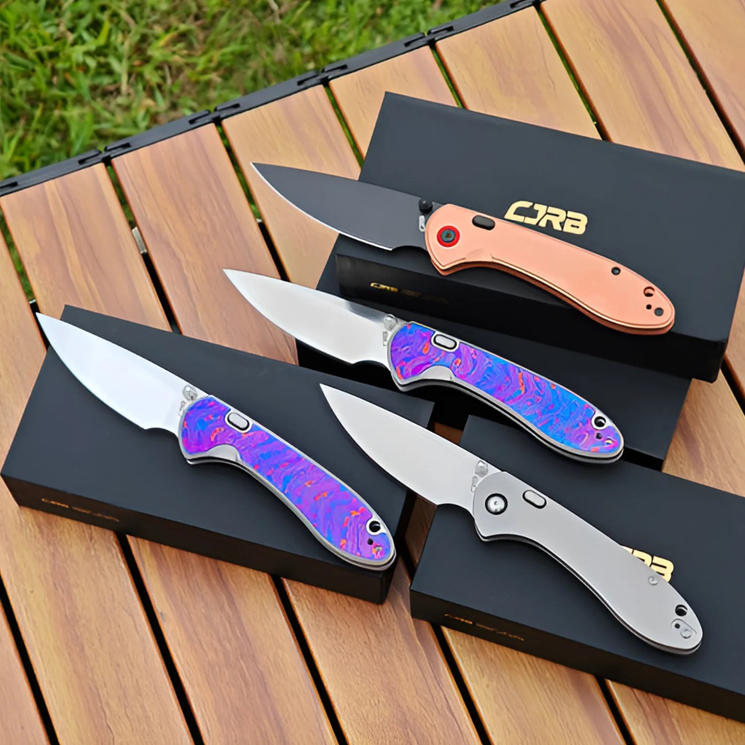 Four folding knives with different handle colors—copper, silver, and two iridescent purple—displayed on black boxes on a wooden surface.