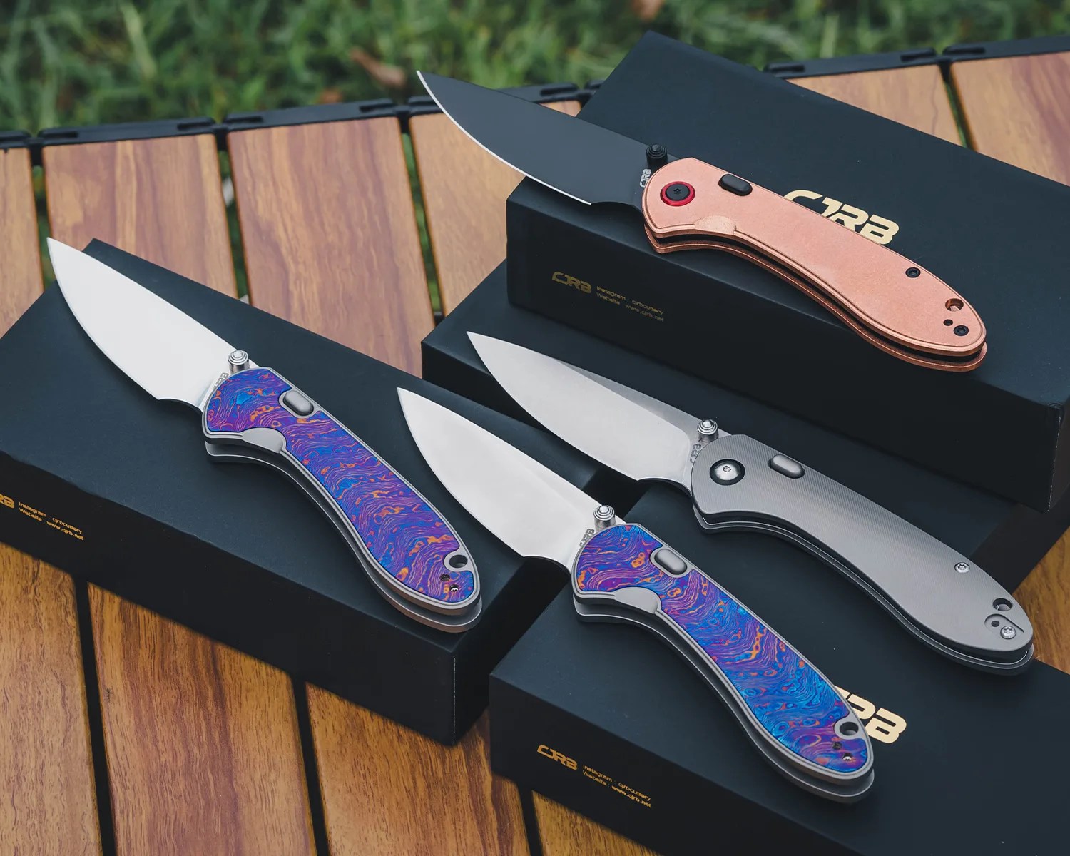 Four folding knives with different handle designs, including copper, gray, and blue-purple patterned handles, displayed on black CRB boxes on a wooden surface.