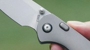 Close-up of a silver folding knife blade and handle with a thumb stud and button held in a hand.