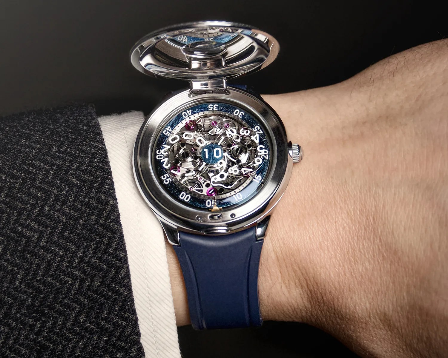 Silver mechanical wristwatch with open face showing intricate gears and a blue strap worn on a wrist.