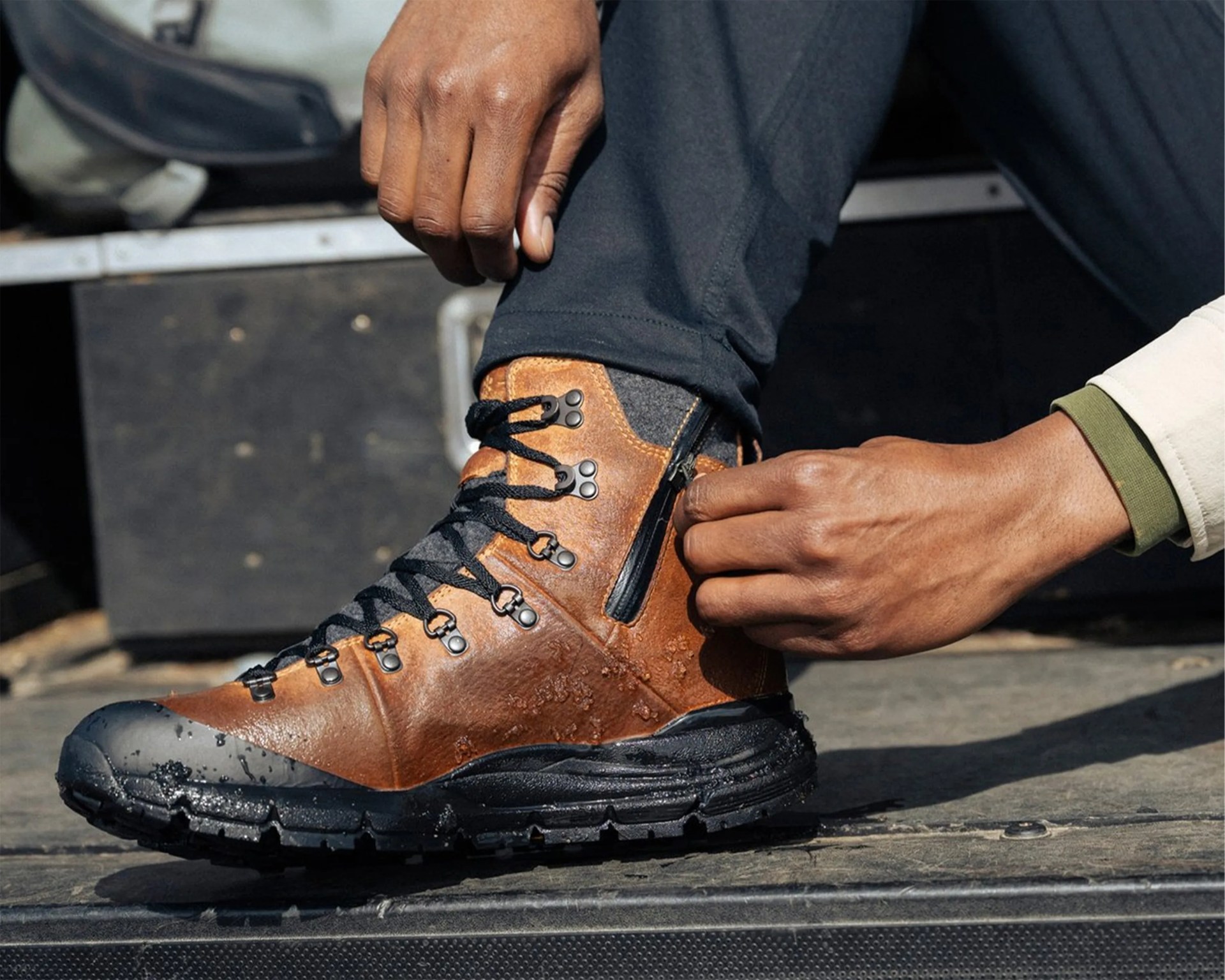 Brown leather hiking boot with black sole and laces being zipped up on a foot wearing black pants.