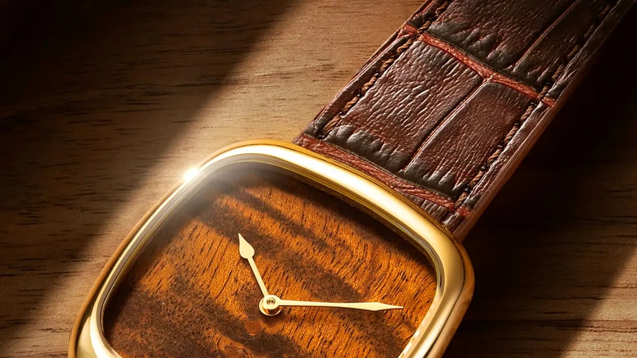 Square wristwatch with a gold-tone frame, wooden dial, and brown leather strap on a wooden surface.