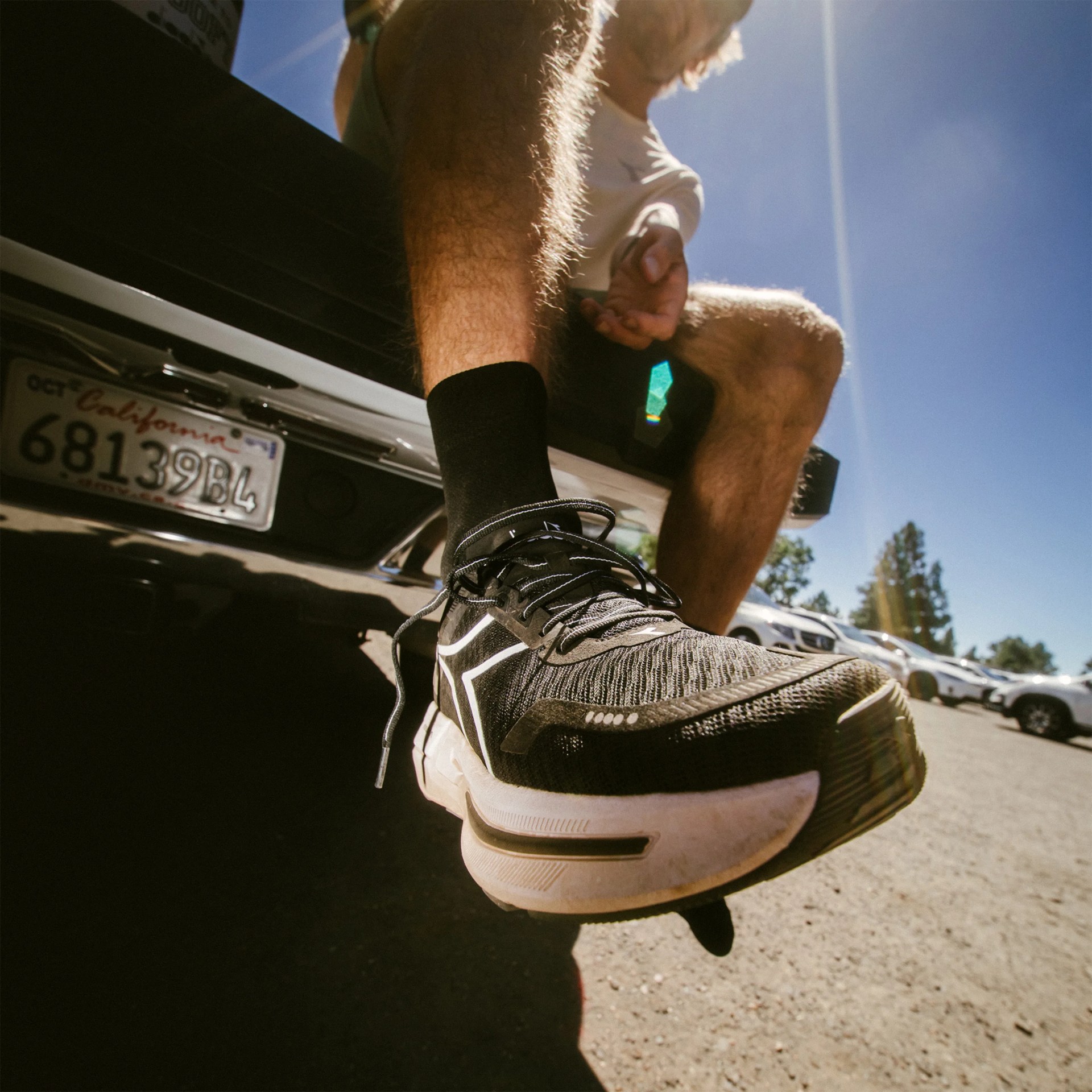 Black and white running shoe worn by a person sitting on the back of a vehicle with a California license plate.