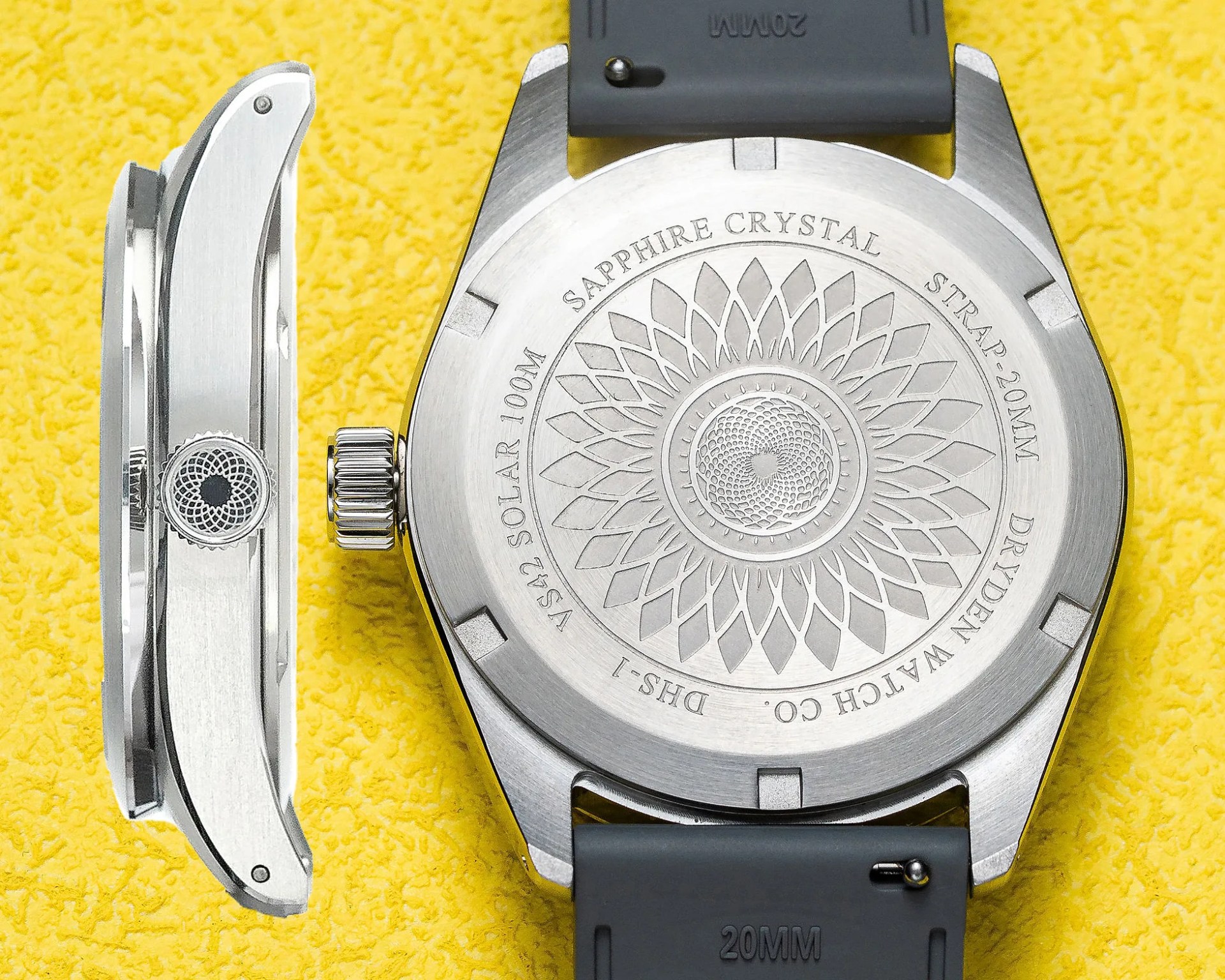 Back of a stainless steel wristwatch with engraved floral pattern and black rubber strap on a yellow textured surface.