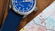 Close-up of a blue Heartlander watch with a blue strap resting on a brown textured surface next to a folded topographic map.