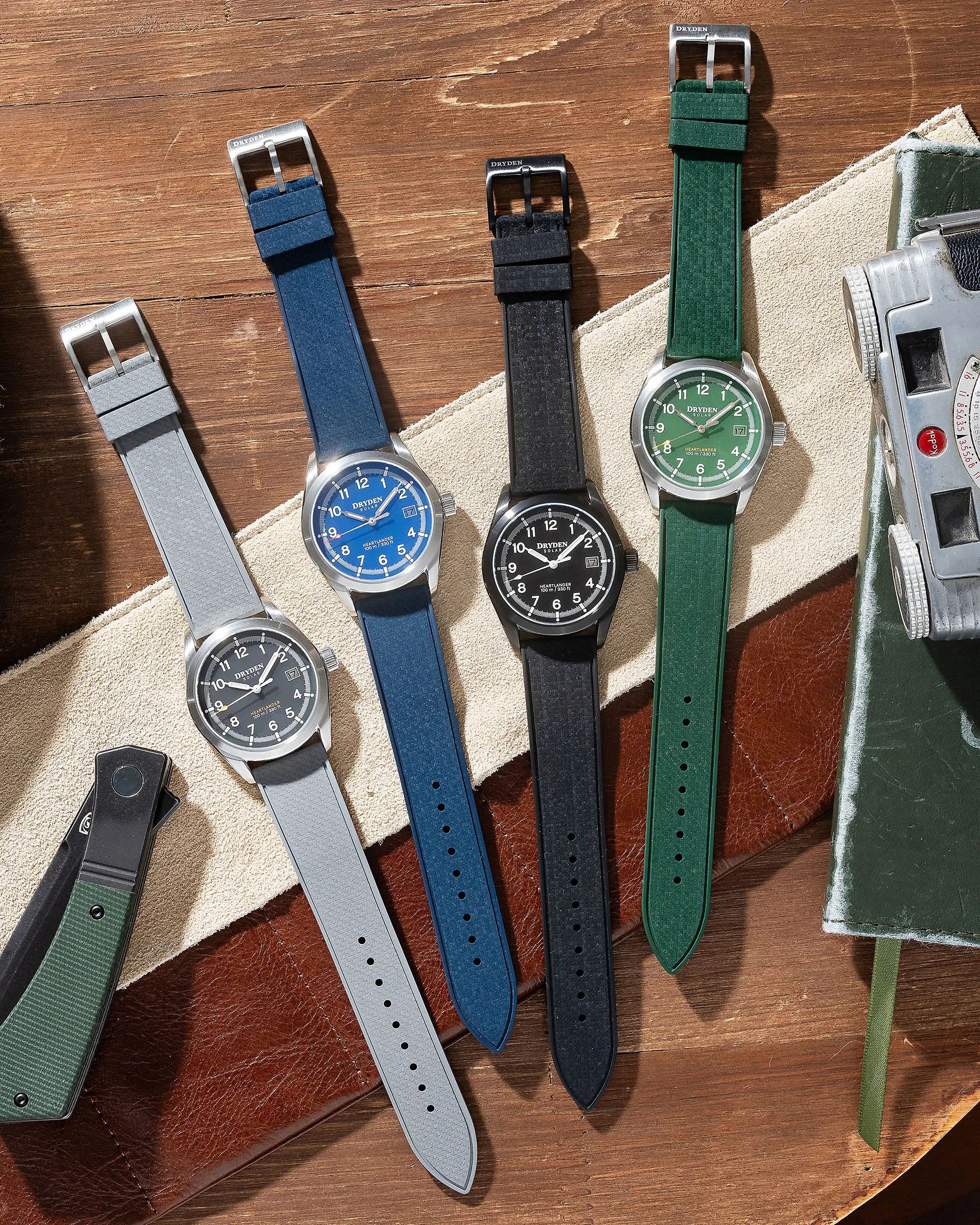 Four wristwatches with textured straps in gray, blue, black, and green, each with a round face and date display, laid on a wooden surface.