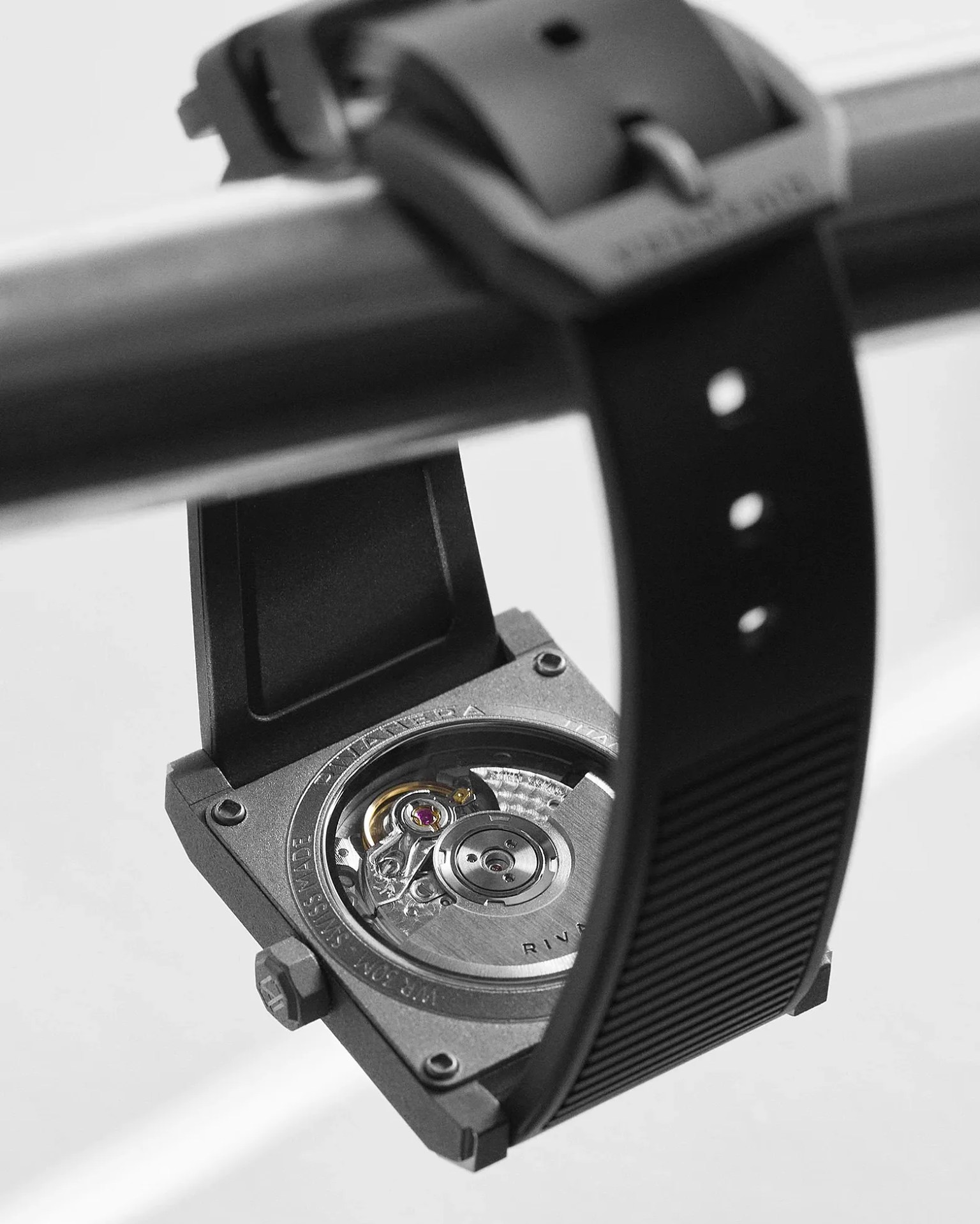 Close-up of the back of a square mechanical wristwatch with a black rubber strap hanging on a metal bar.