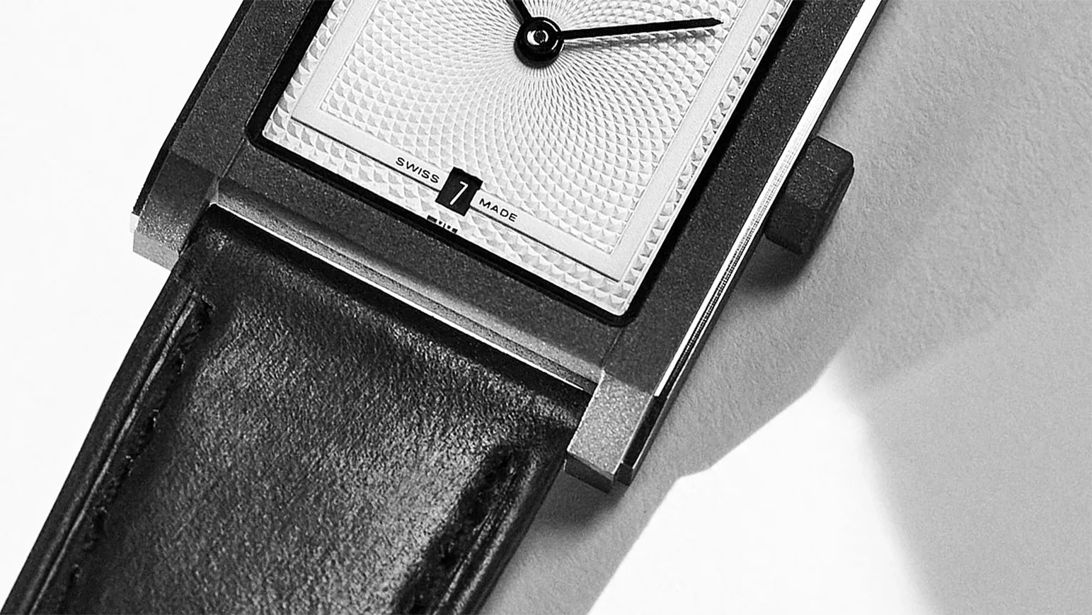 Close-up of a rectangular watch face with textured dial, black hands, date window showing "1," and black leather strap.