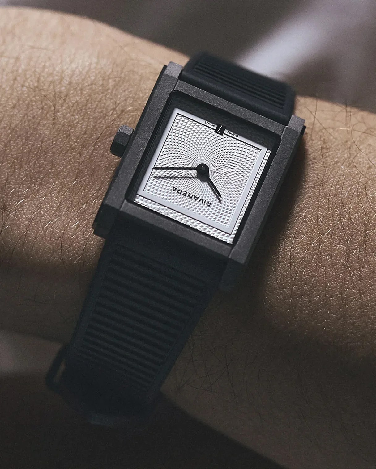 Square black wristwatch with textured silver dial and black rubber strap worn on a wrist.