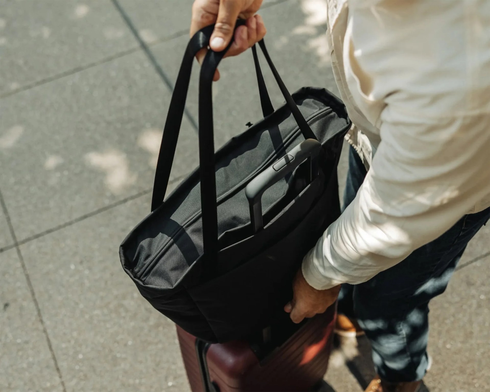 Black tote bag with long handles attached to the handle of a maroon rolling suitcase, held by a person in a light jacket.