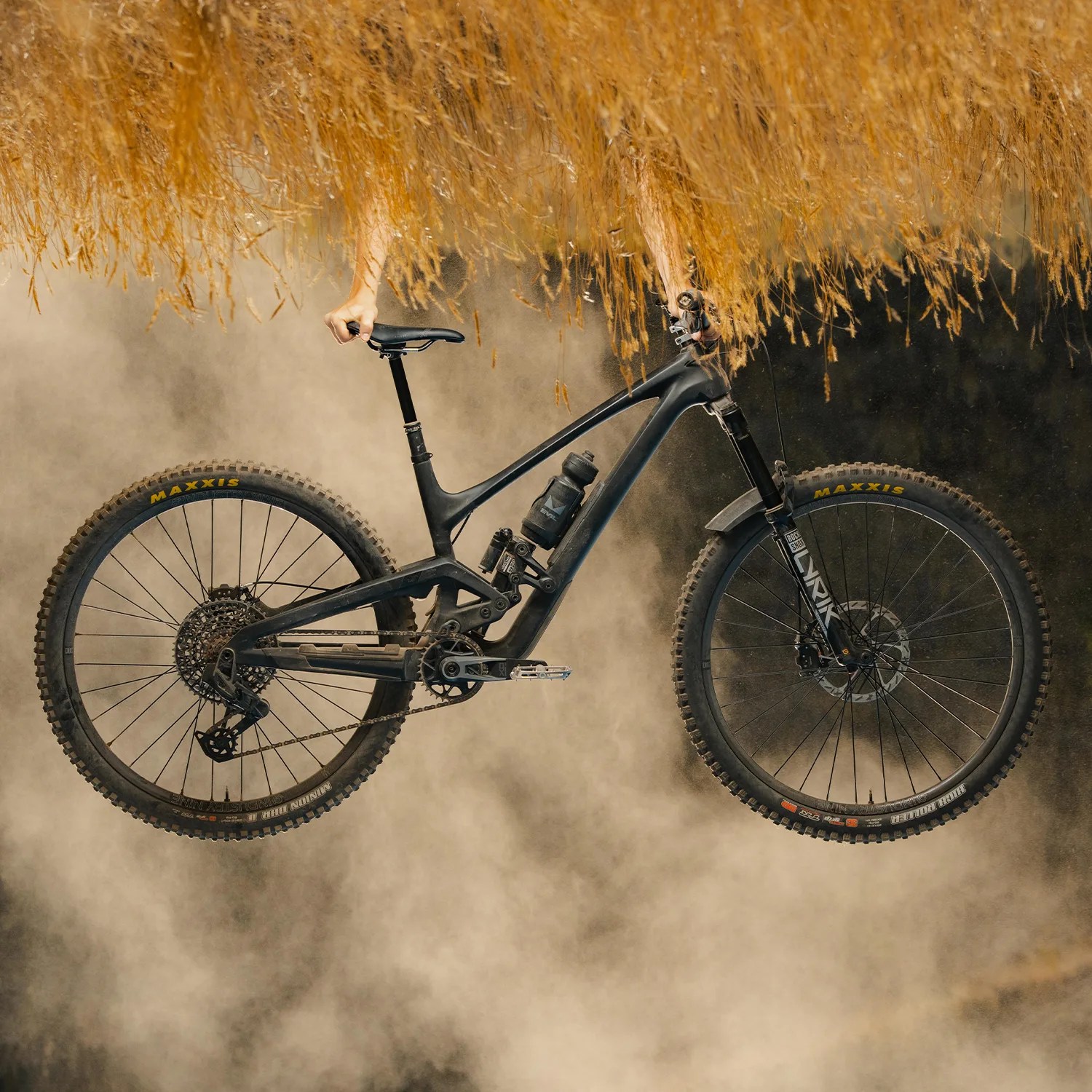 Black full-suspension mountain bike with Maxxis tires held upside down in a dusty outdoor setting.