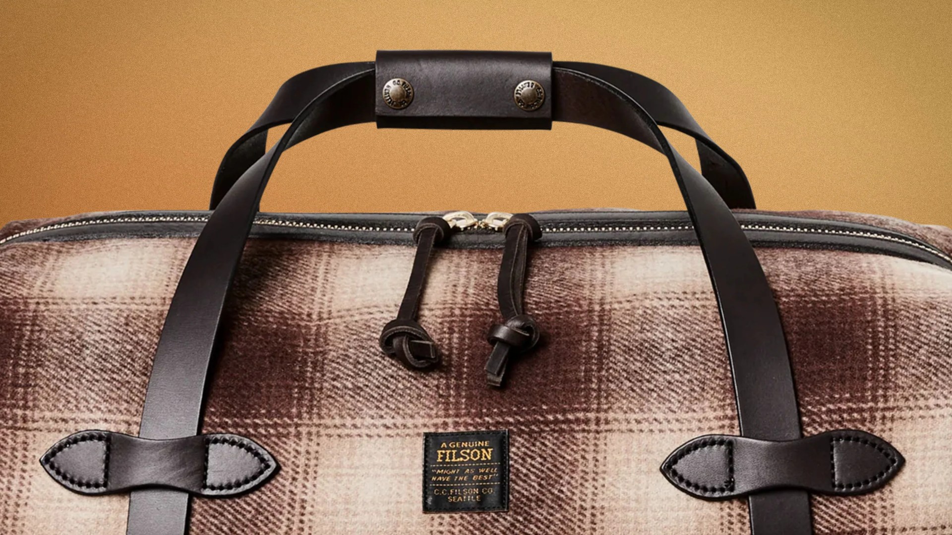 Brown and beige plaid fabric duffel bag with black leather handles and zipper pulls, featuring a Filson label.