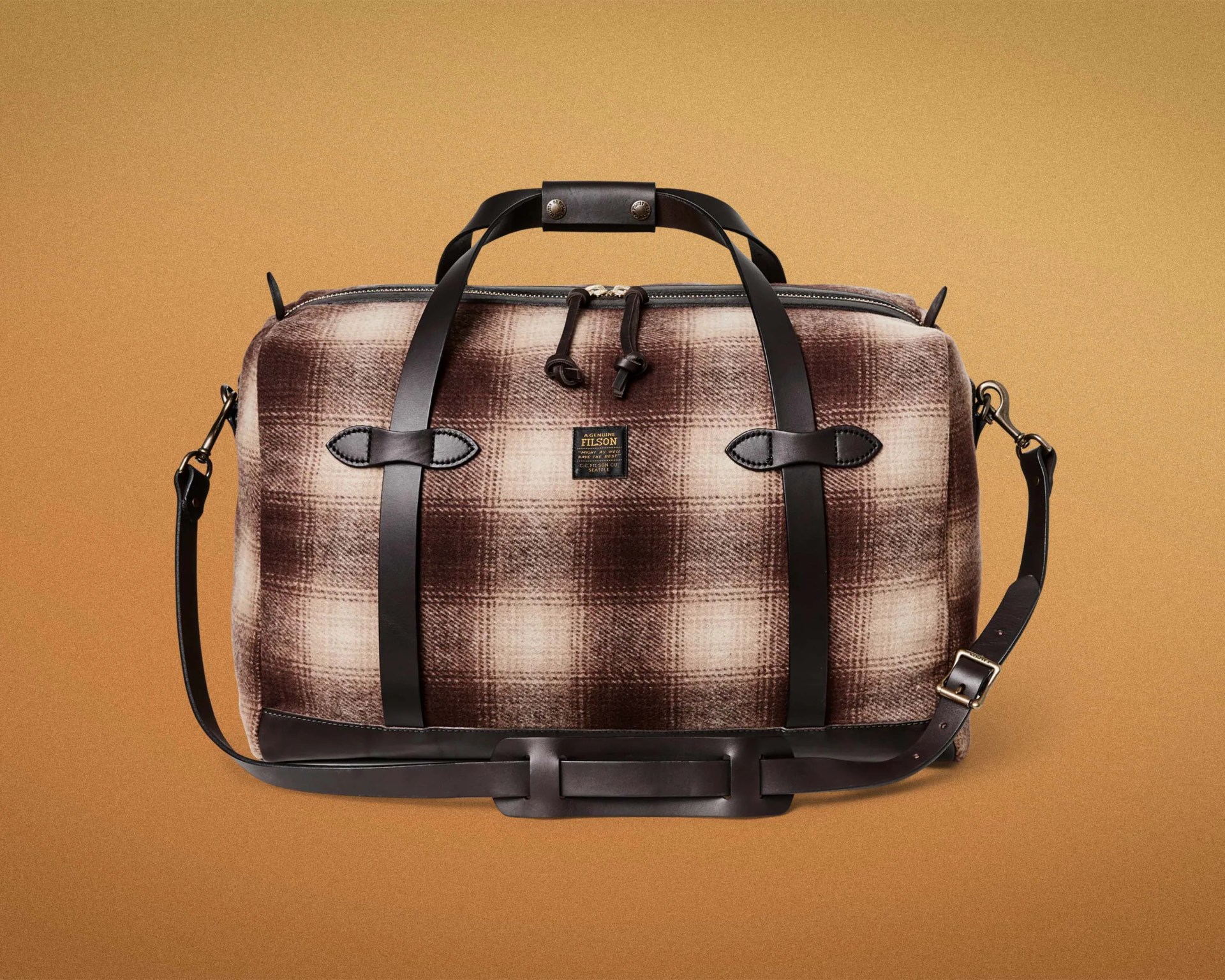 Brown and beige plaid duffel bag with black leather handles and adjustable shoulder strap on a tan background.