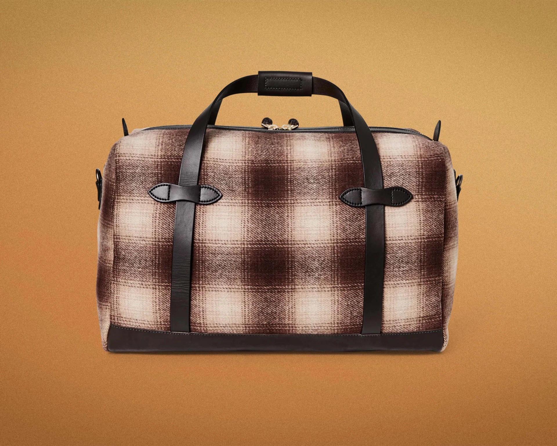 Brown and beige plaid fabric duffel bag with black leather handles and trim on a tan background.