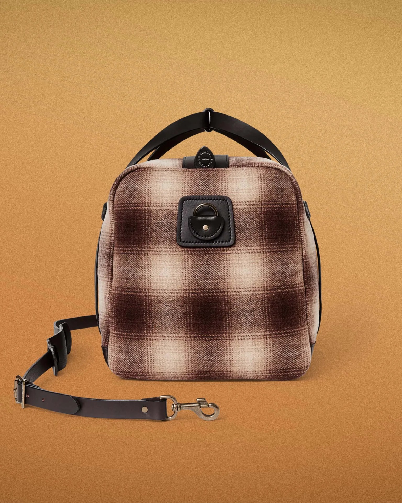 Brown and beige plaid fabric backpack with black leather straps and metal clasp on a brown background.