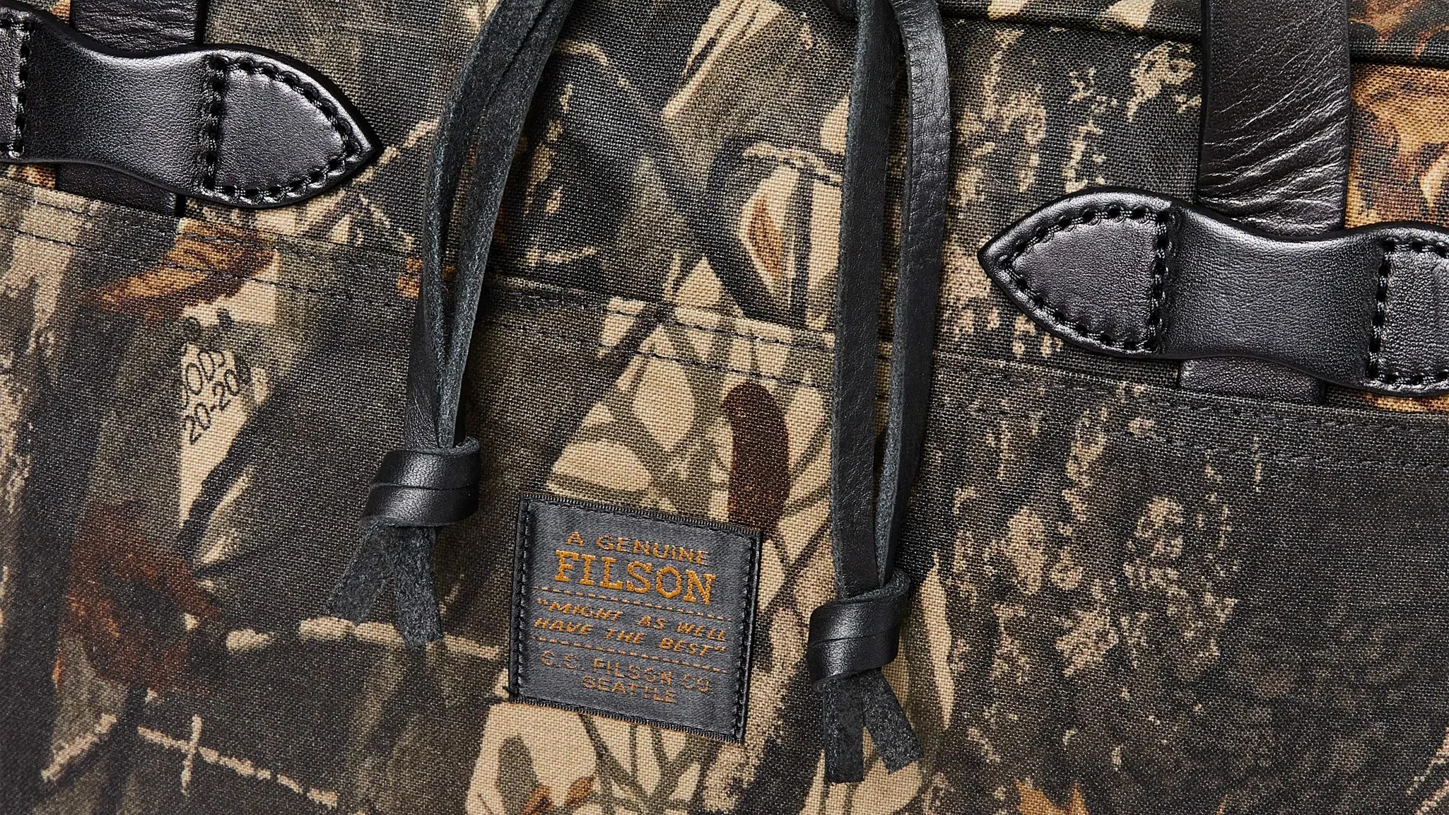 Close-up of a camouflage fabric bag with black leather straps and a label reading "A Genuine Filson.