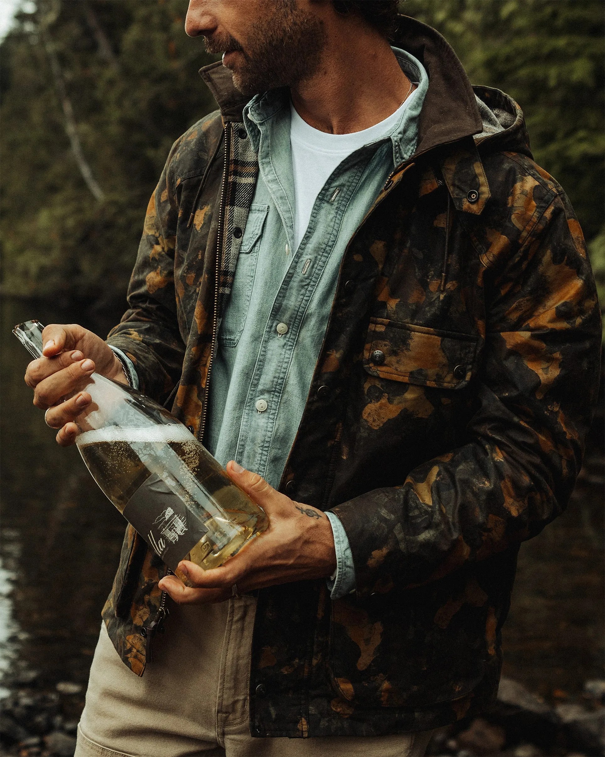 Man wearing a dark brown and orange camo jacket over a light blue denim shirt holding a large clear bottle with a black label.
