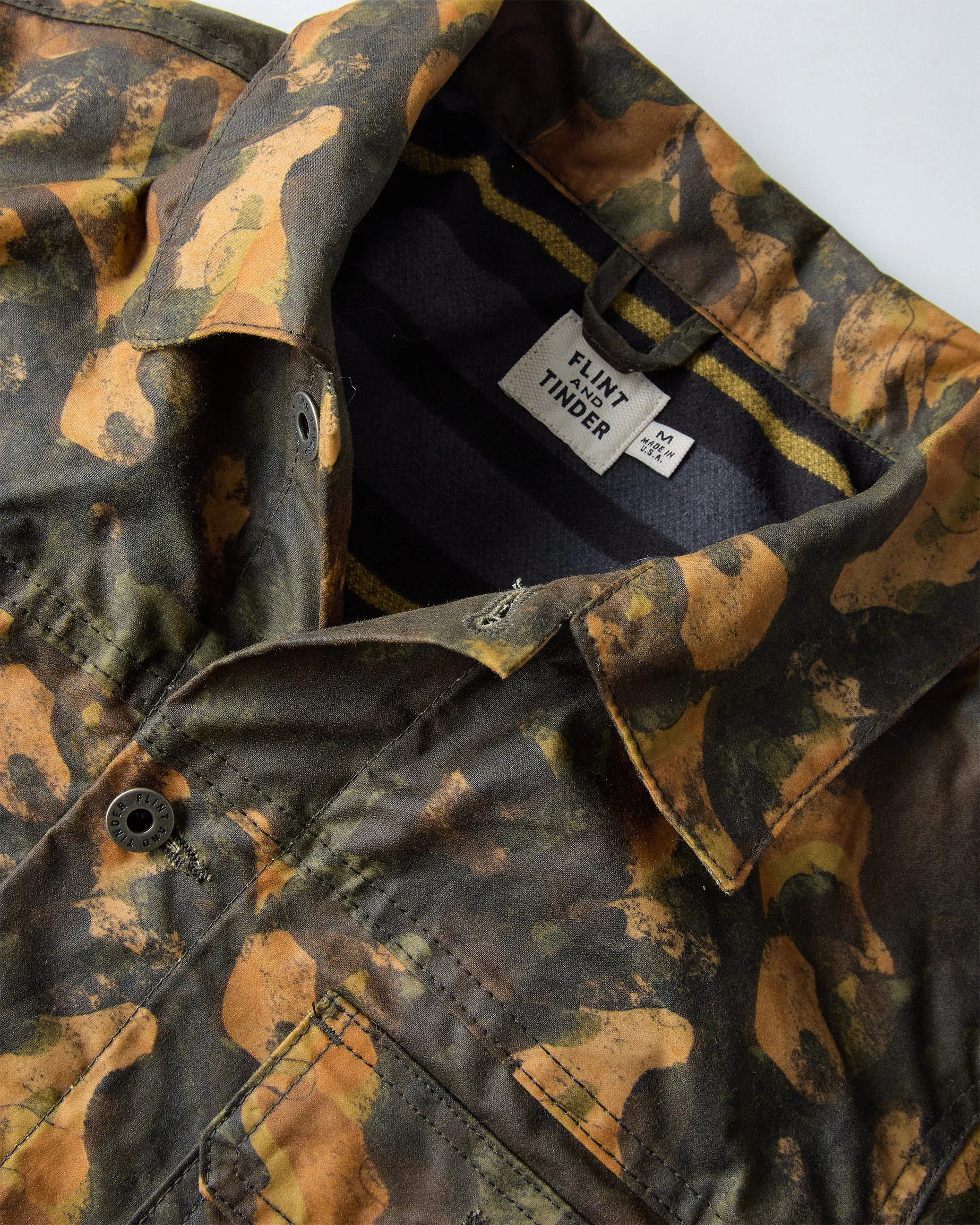 Close-up of a camo-patterned jacket collar with a "Flint and Tinder" label and black button.