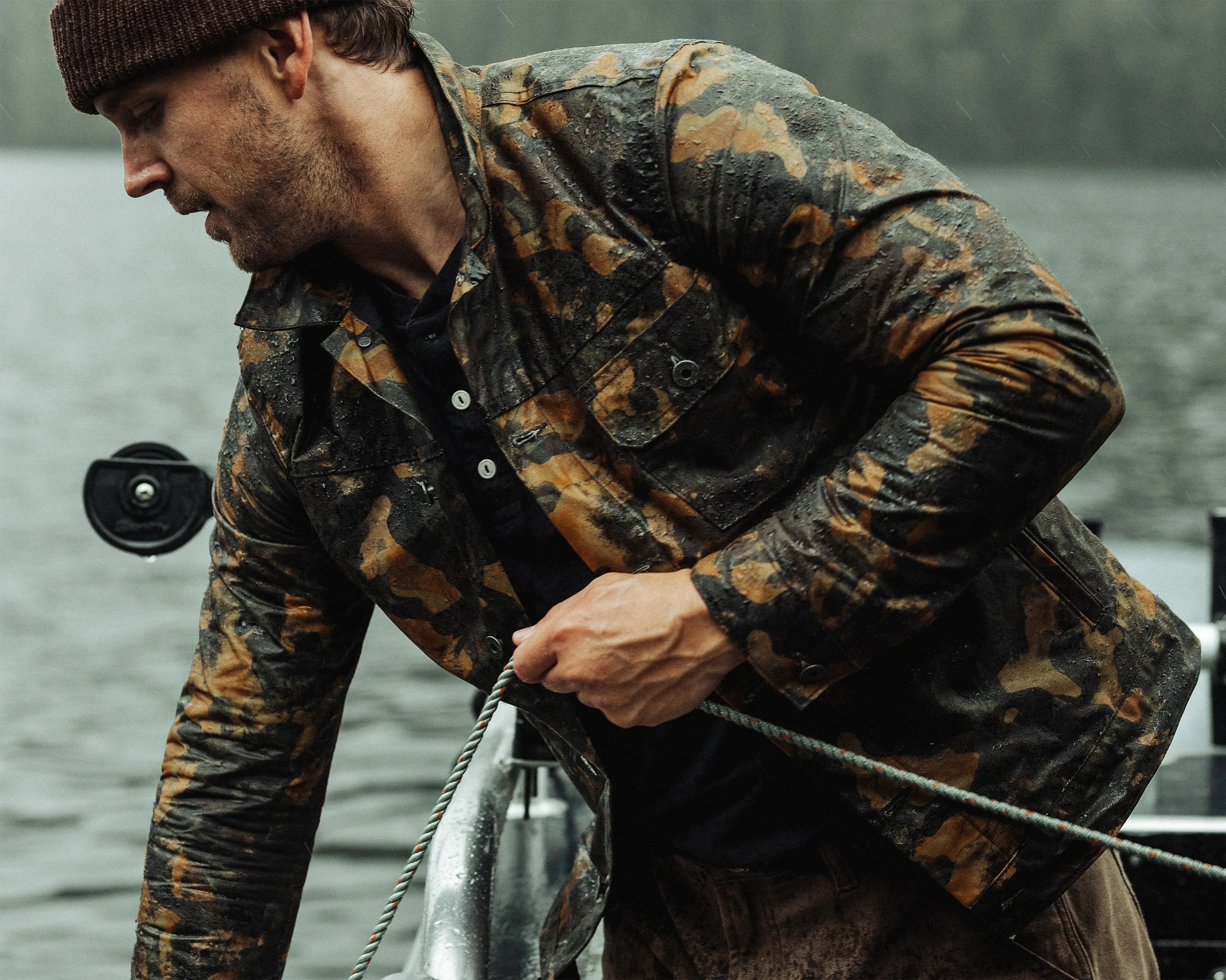 Man wearing a wet brown and black camouflage jacket and brown knit hat holding a rope on a boat.