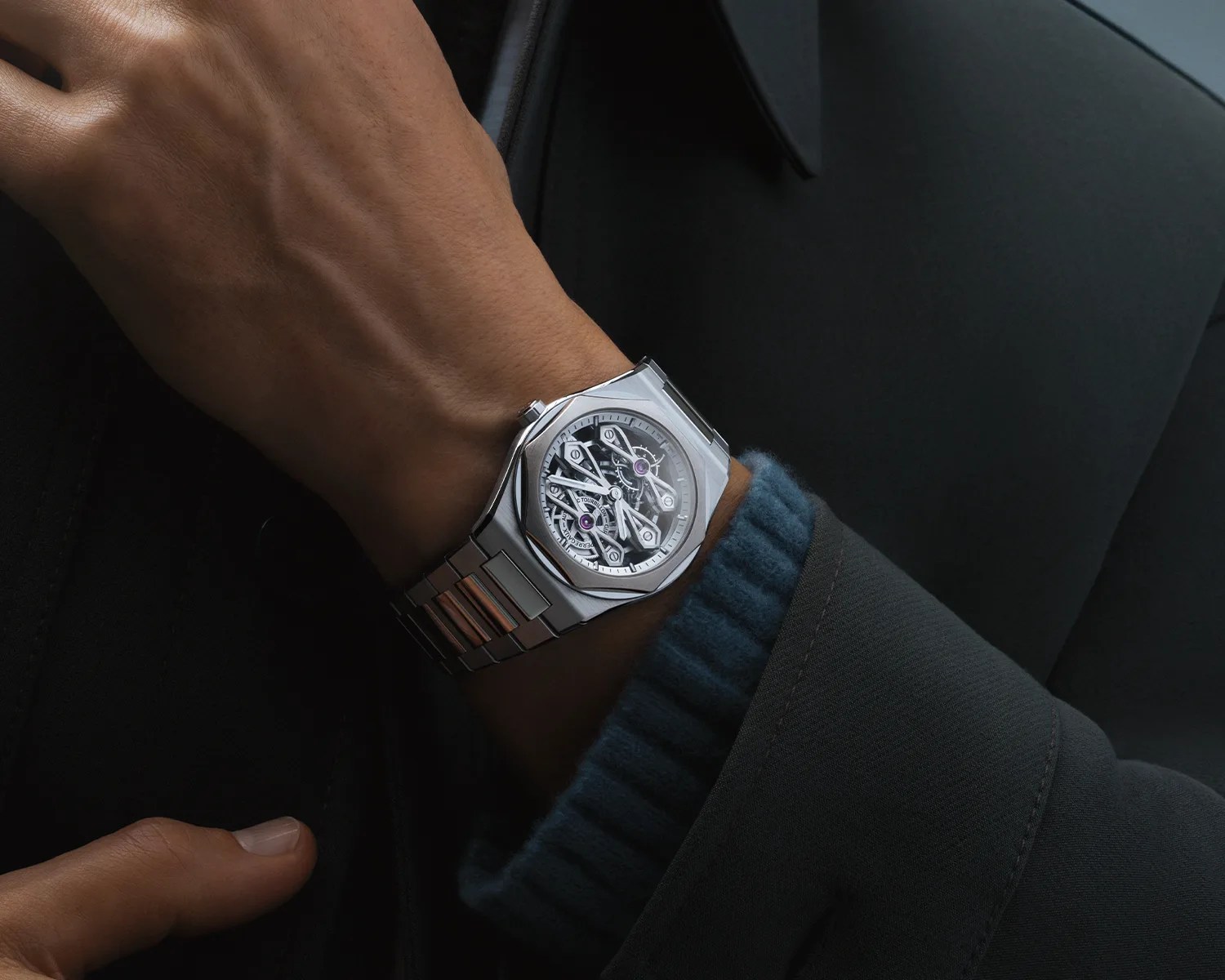 Silver metal wristwatch with a skeleton dial showing internal gears, worn on a wrist with a dark jacket and blue cuff.
