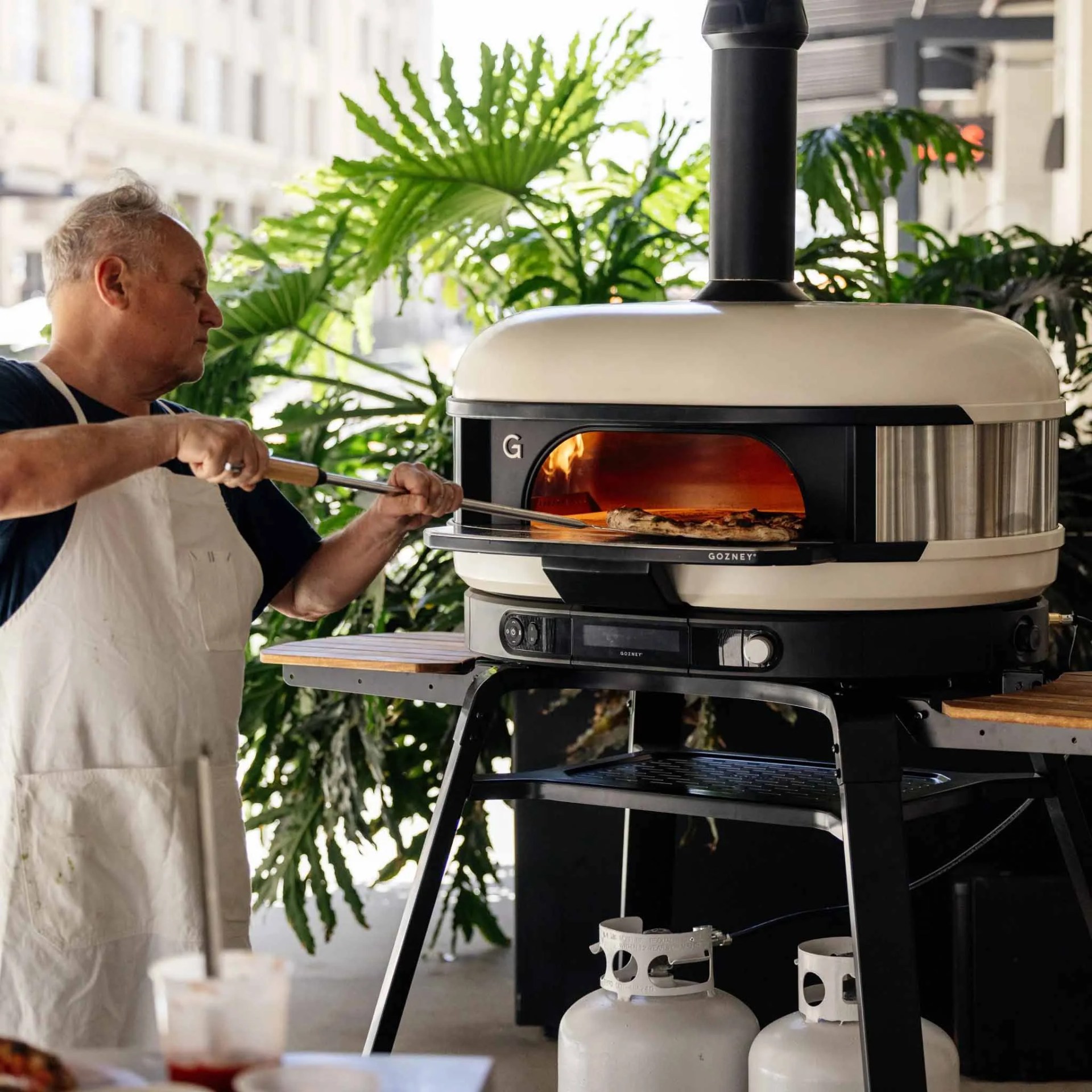 Man in white apron using a Gozney outdoor pizza oven with a pizza peel and pizza inside.