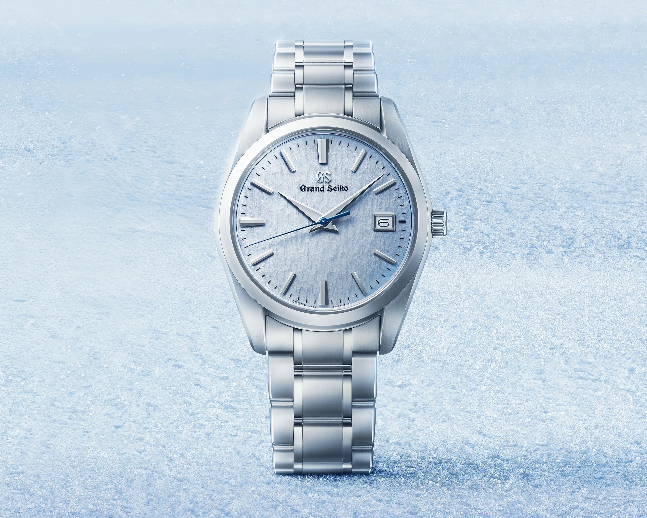 Silver Grand Seiko wristwatch with textured light blue dial and date window on icy blue background.