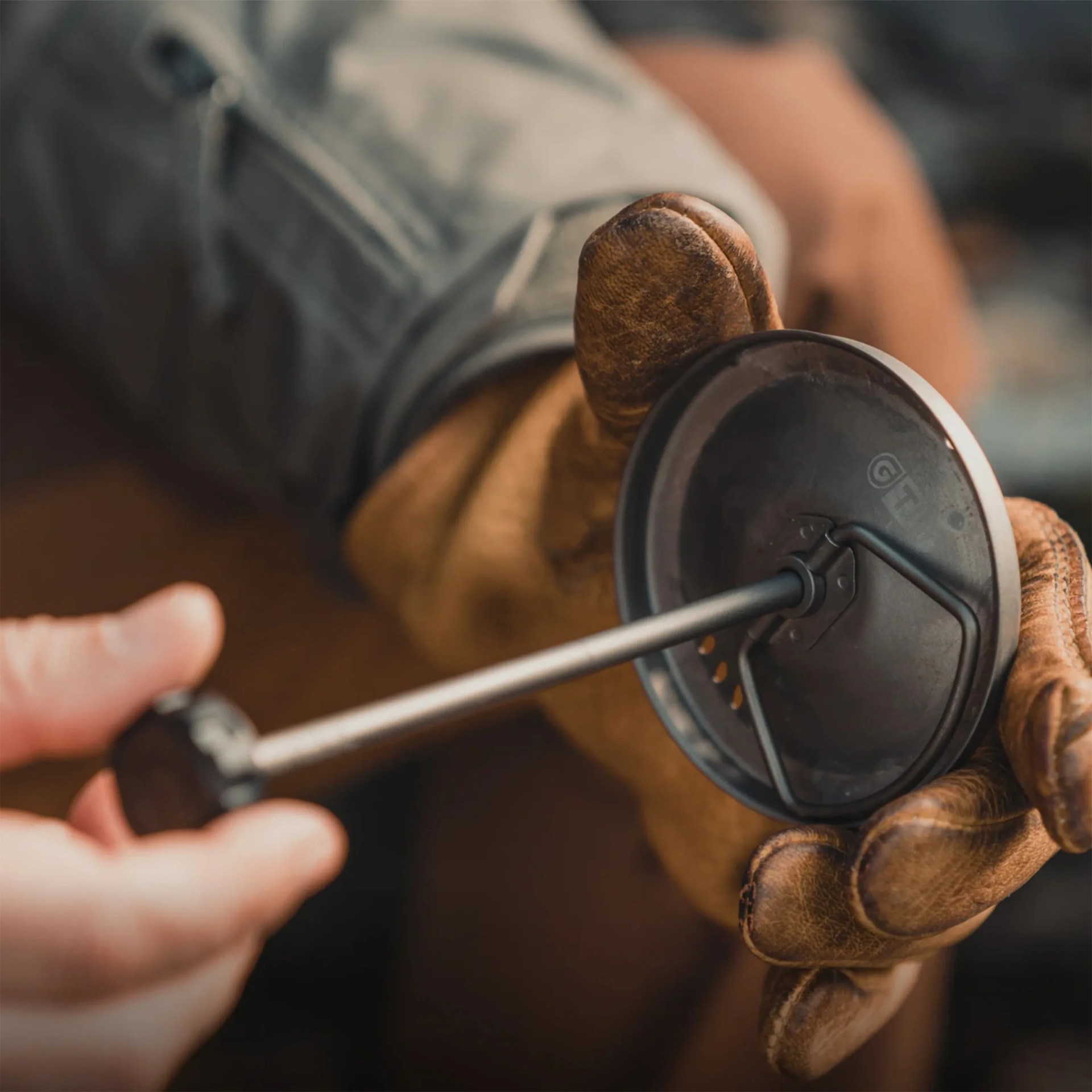 Person wearing brown leather gloves holding a black metal tool with a round flat end and a handle.