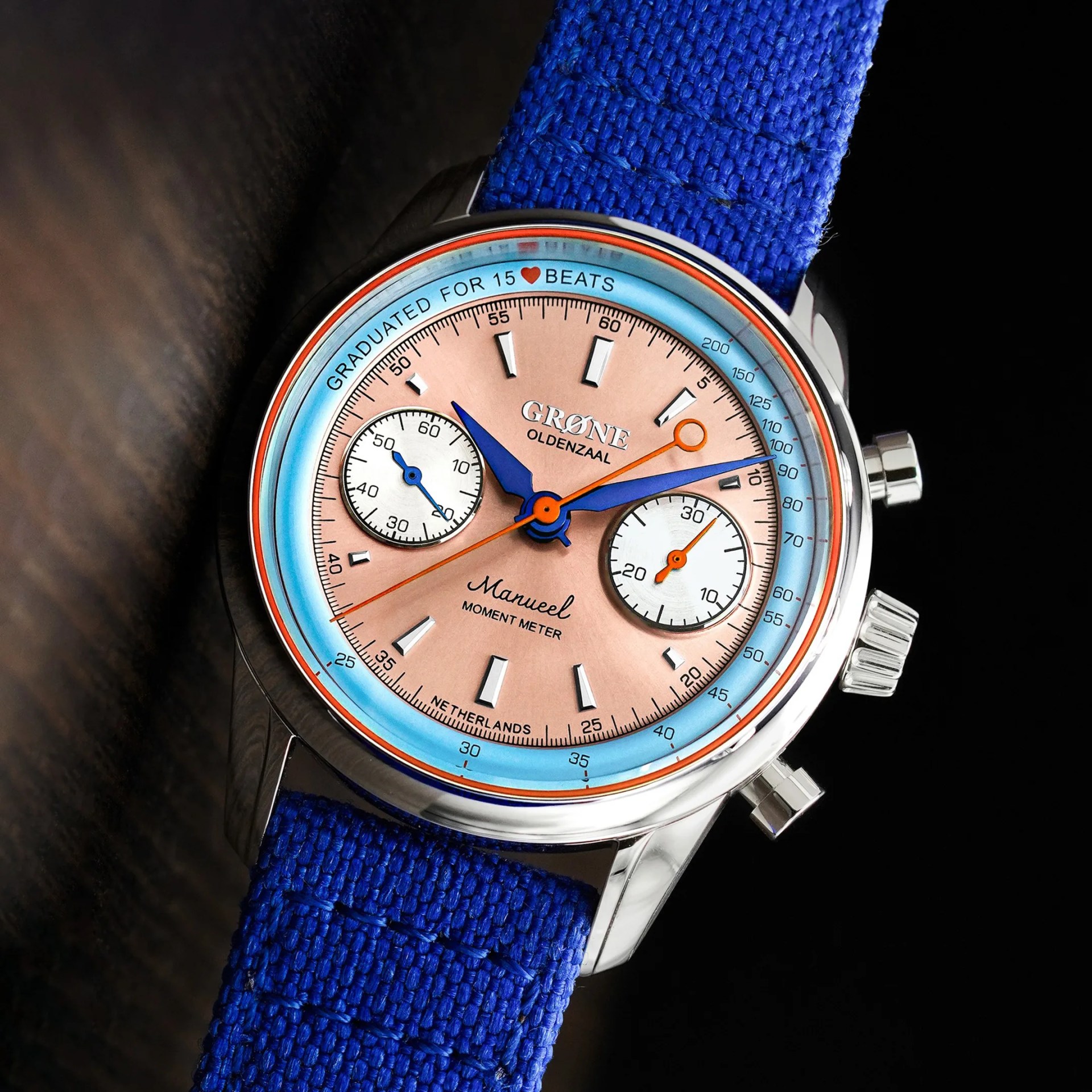 Silver wristwatch with a pink dial, blue and orange hands, two white subdials, and a blue fabric strap.