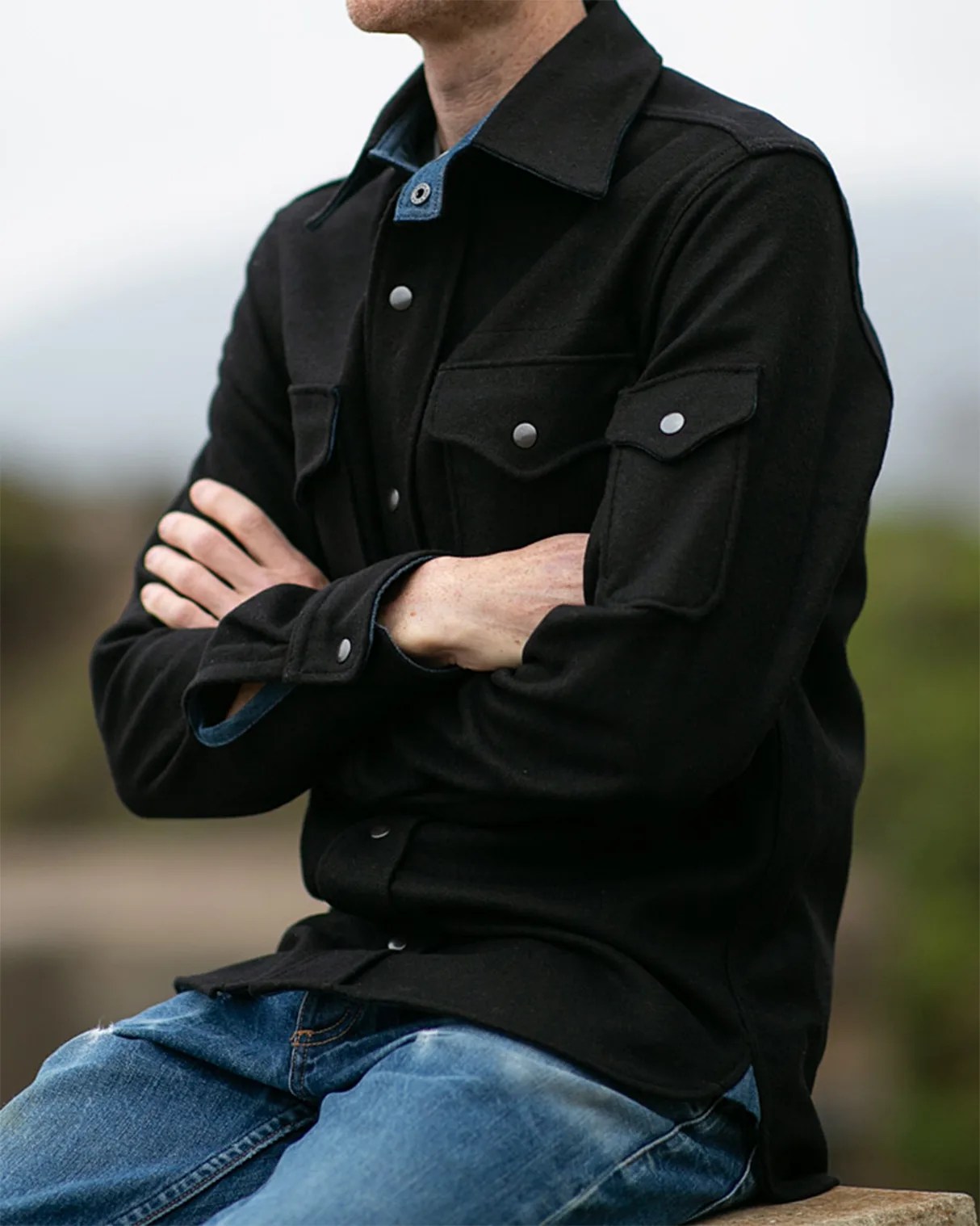 Black button-up jacket with flap pockets on chest and sleeve worn over blue denim jeans.