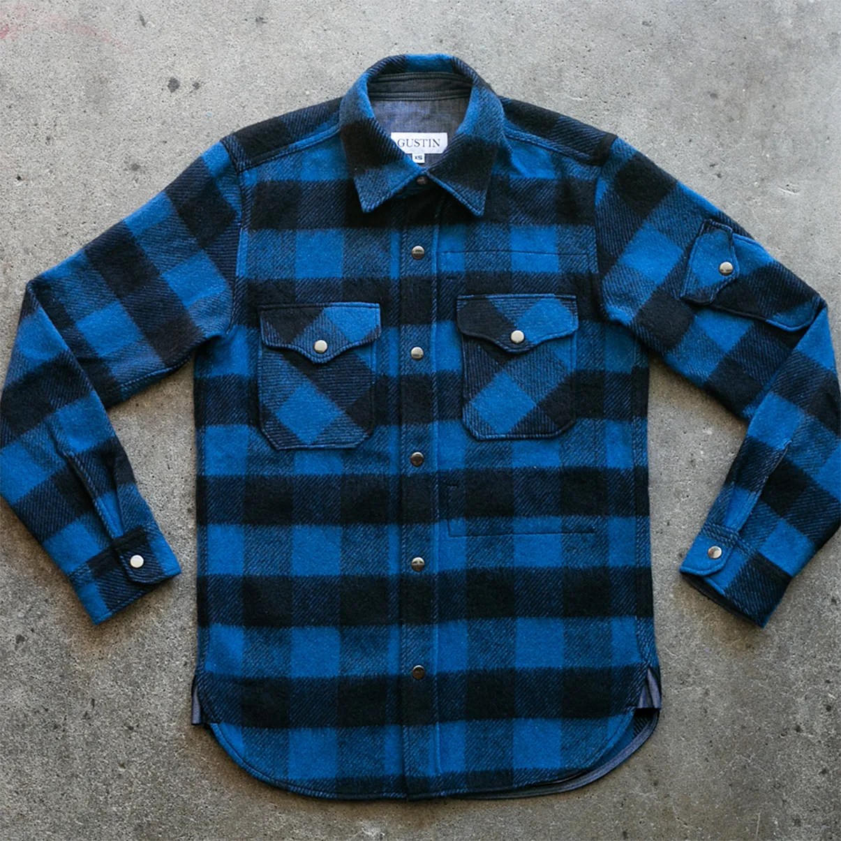 Blue and black checkered flannel shirt with snap buttons and two chest pockets laid flat on concrete.