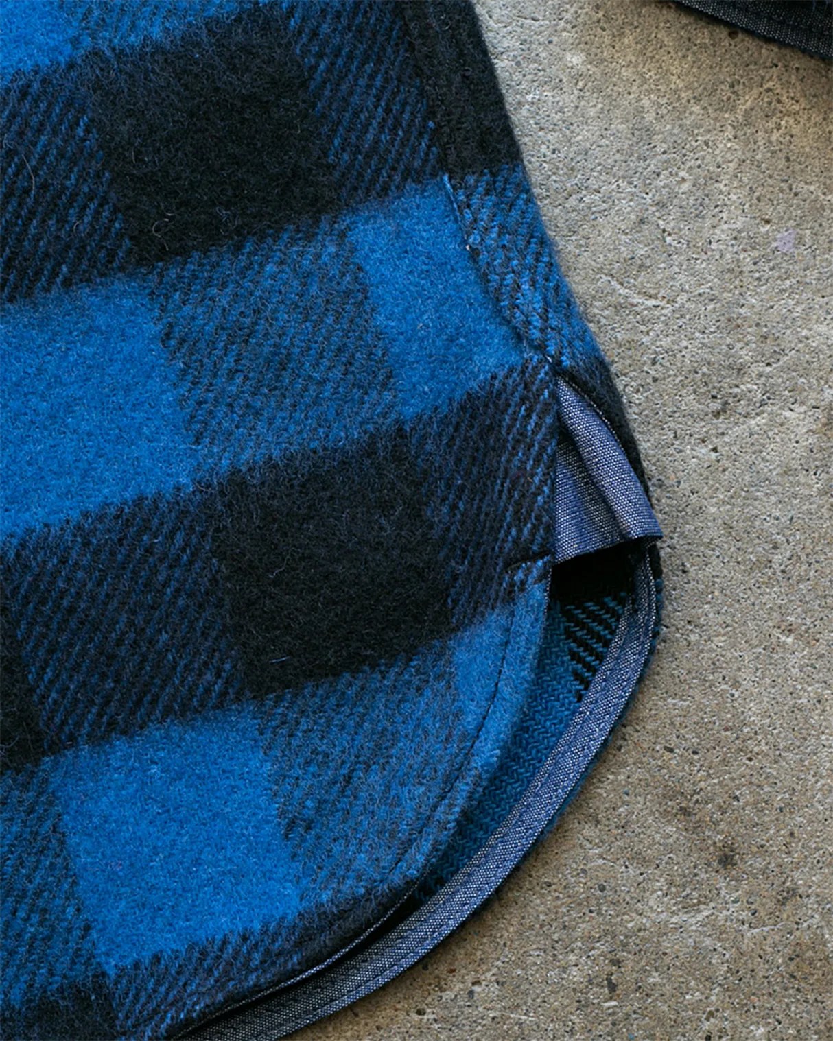 Close-up of blue and black checkered wool fabric with a curved hem and blue inner lining on a concrete surface.
