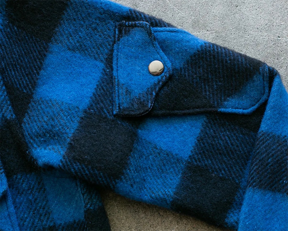Blue and black checkered wool jacket sleeve with a flap pocket and silver snap button on a concrete surface.