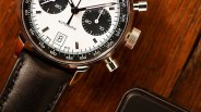 Close-up of a silver chronograph watch with a white dial, black subdials, and a black leather strap on a wooden surface.