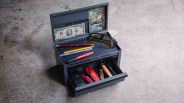 Small gray U.S. General metal toolbox with open top and drawer containing tools, pencils, and sunglasses on a concrete floor.