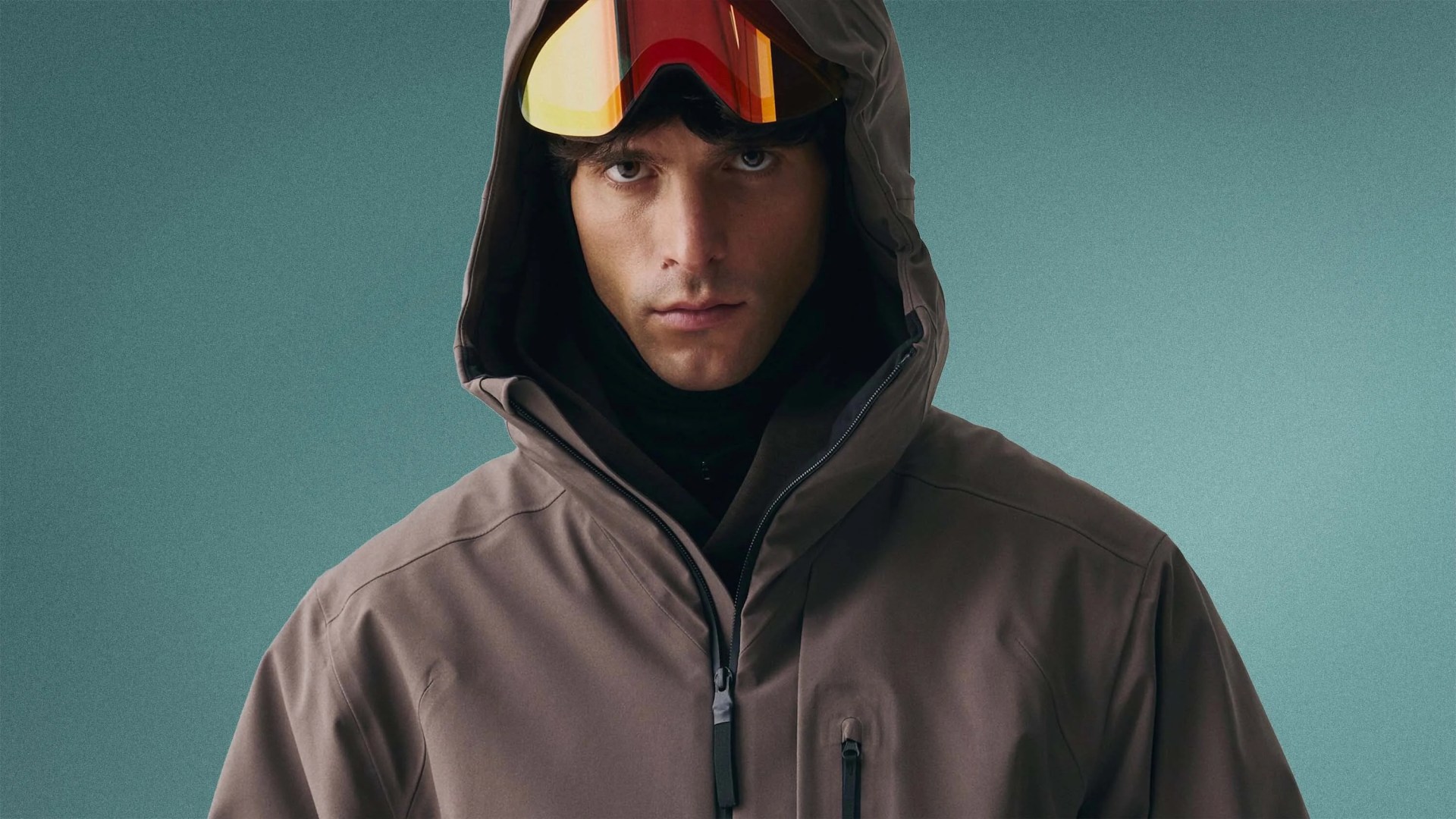 Man wearing a brown hooded jacket with a front zipper and orange-tinted ski goggles on a teal background.