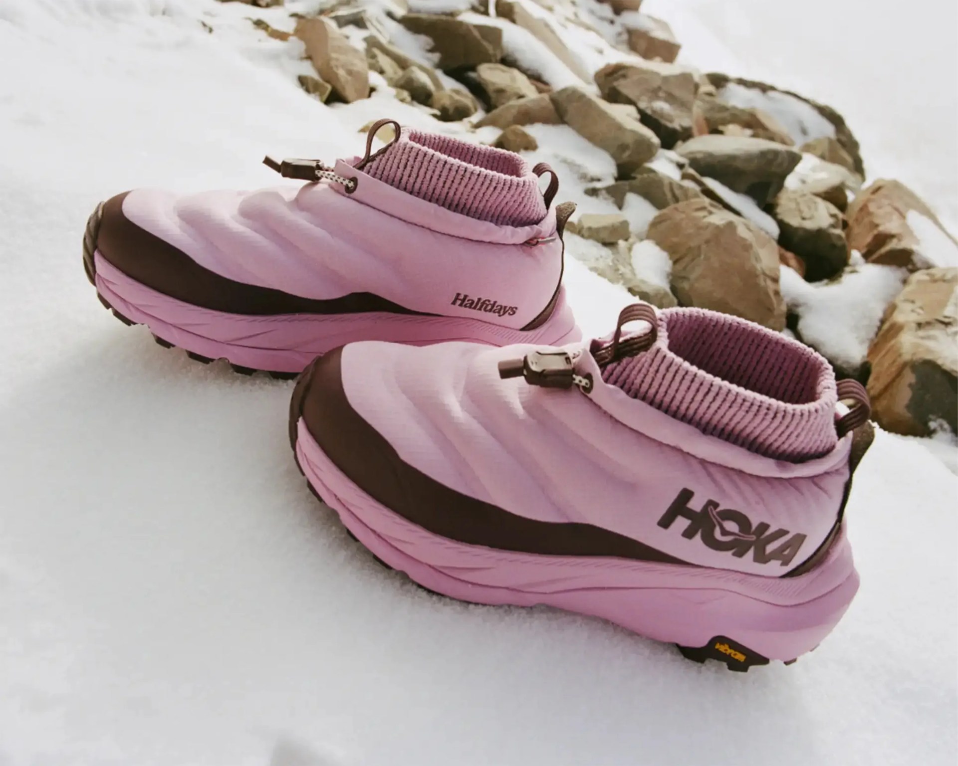 Pair of pink Hoka Halfdays insulated shoes with brown accents on snow near rocks.