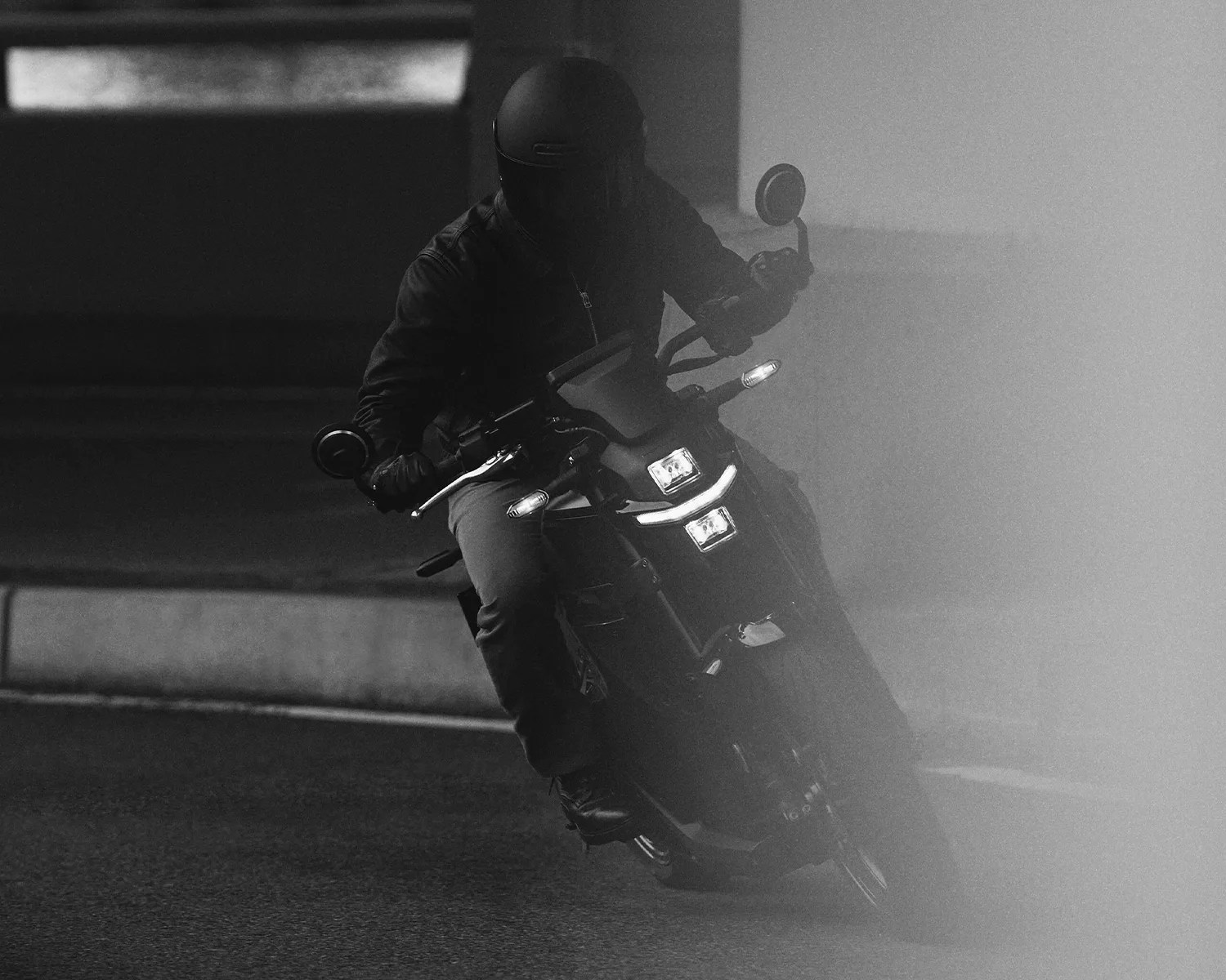 Motorcyclist wearing a helmet and jacket riding a motorcycle with headlights on in low visibility.