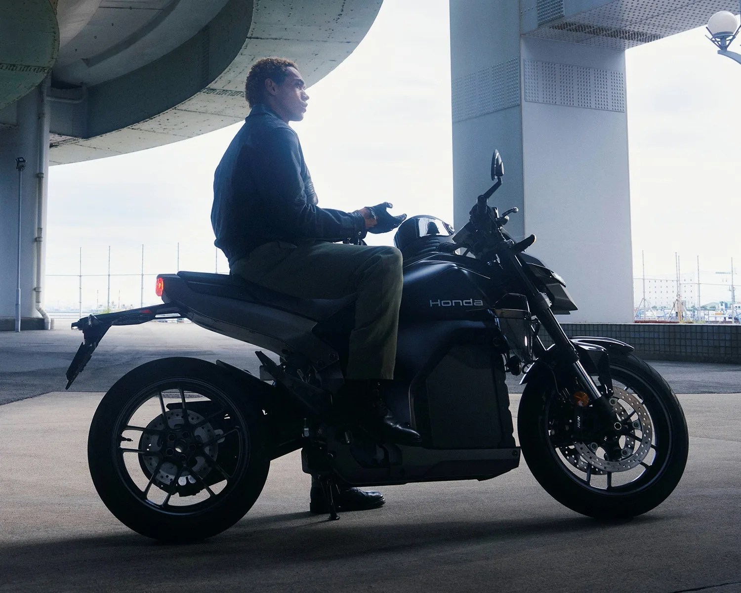 Person sitting on a black Honda motorcycle under an overpass with a helmet on the fuel tank.