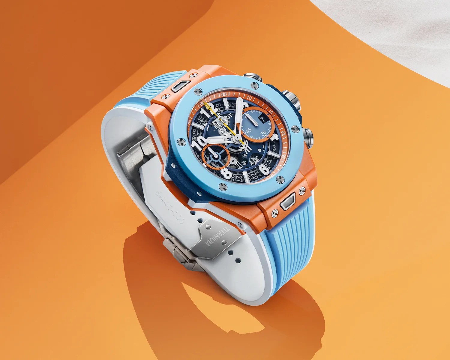 Blue and orange Hublot wristwatch with a skeleton dial and light blue rubber strap on an orange surface.