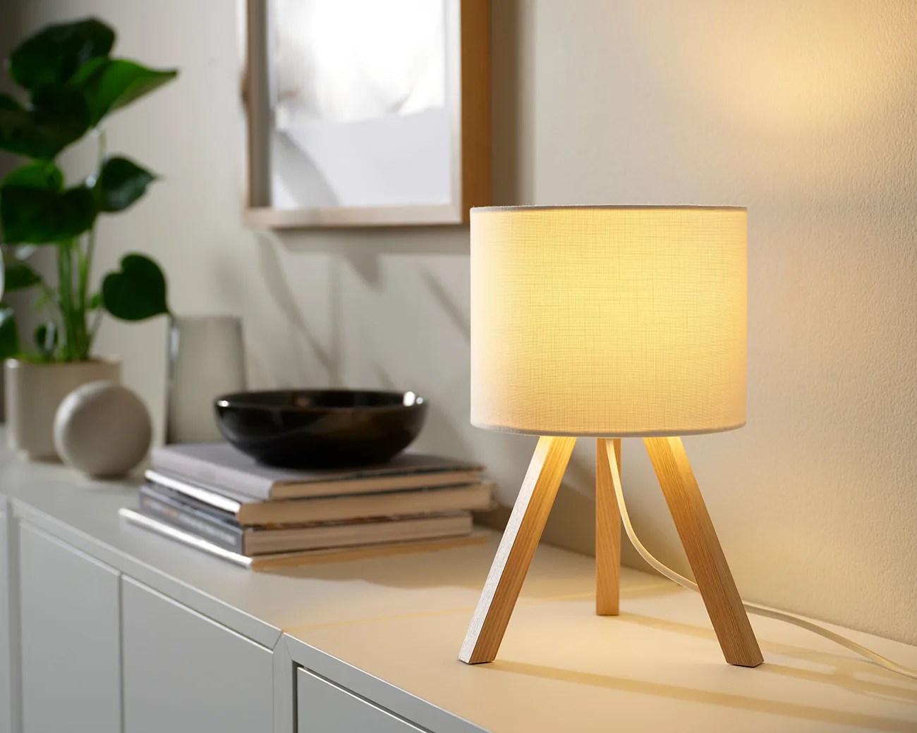 Table lamp with a beige fabric shade and wooden tripod legs on a white cabinet.