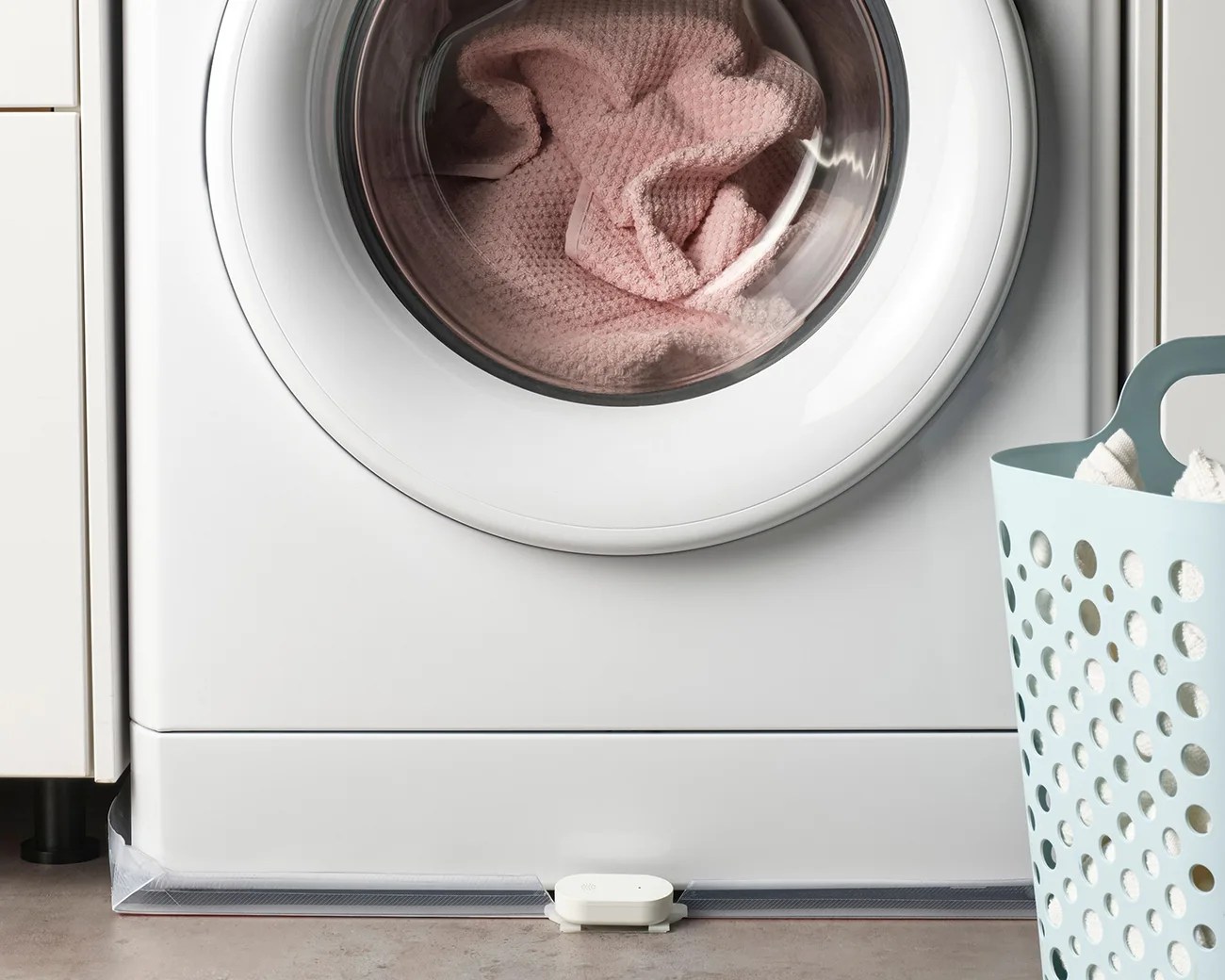 White front-loading washing machine with a pink towel inside and a blue laundry basket filled with white towels nearby.