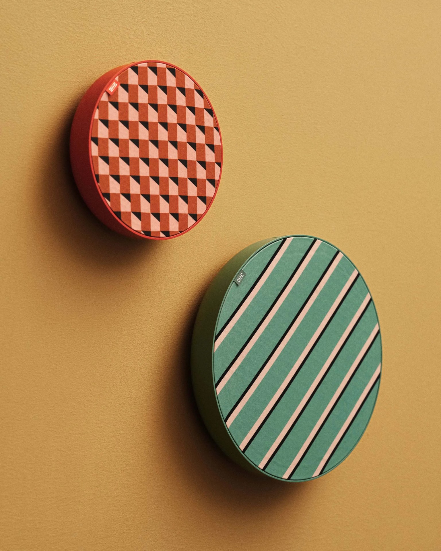 Two round wall-mounted speakers with patterned fabric covers, one in red with geometric shapes and the other in green with diagonal stripes, on a beige wall.