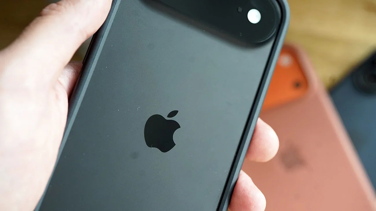 Hand holding a black iPhone with Apple logo and dual camera, with two other iPhones blurred in the background.