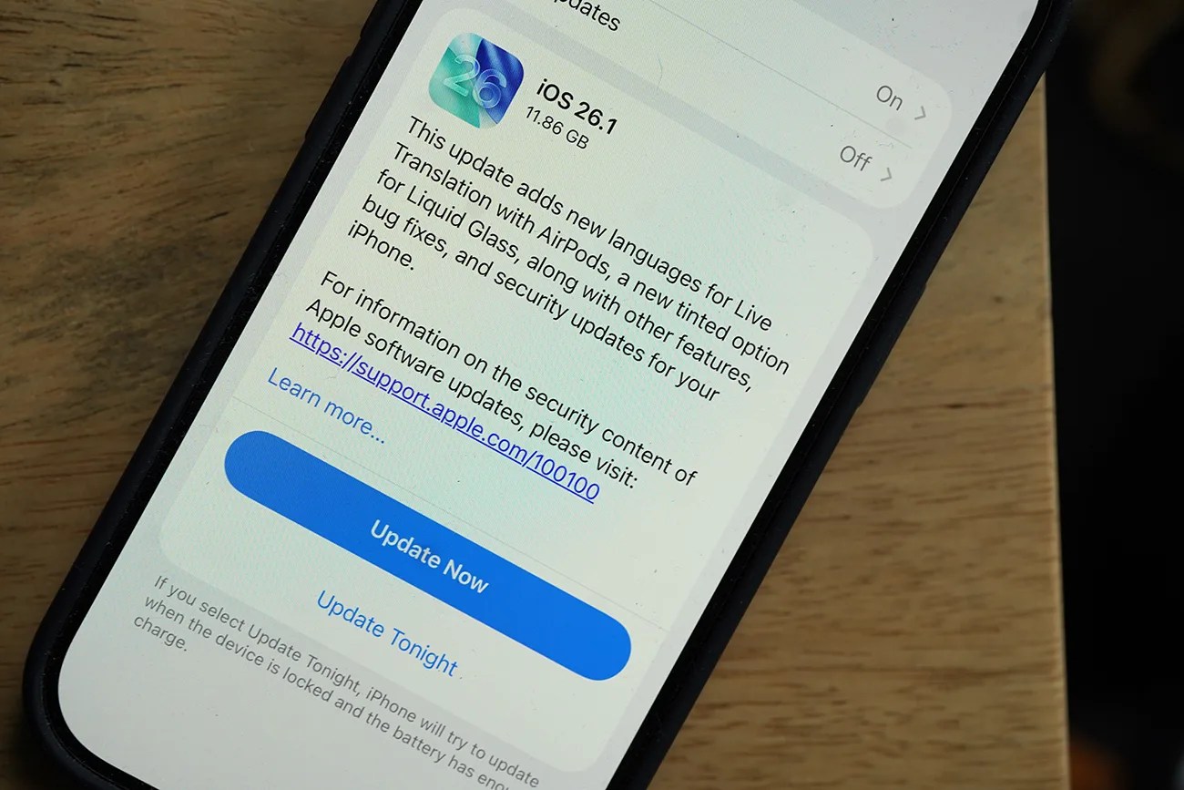 Smartphone screen showing iOS 26.1 update details with an "Update Now" button on a wooden surface.