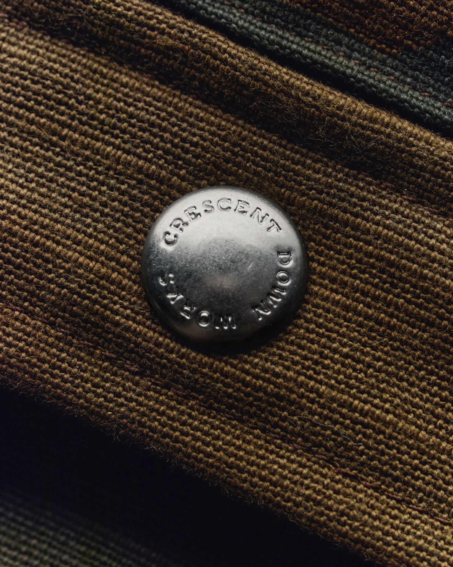 Round silver metal button with "Crescent Down Works" embossed, attached to brown textured fabric.
