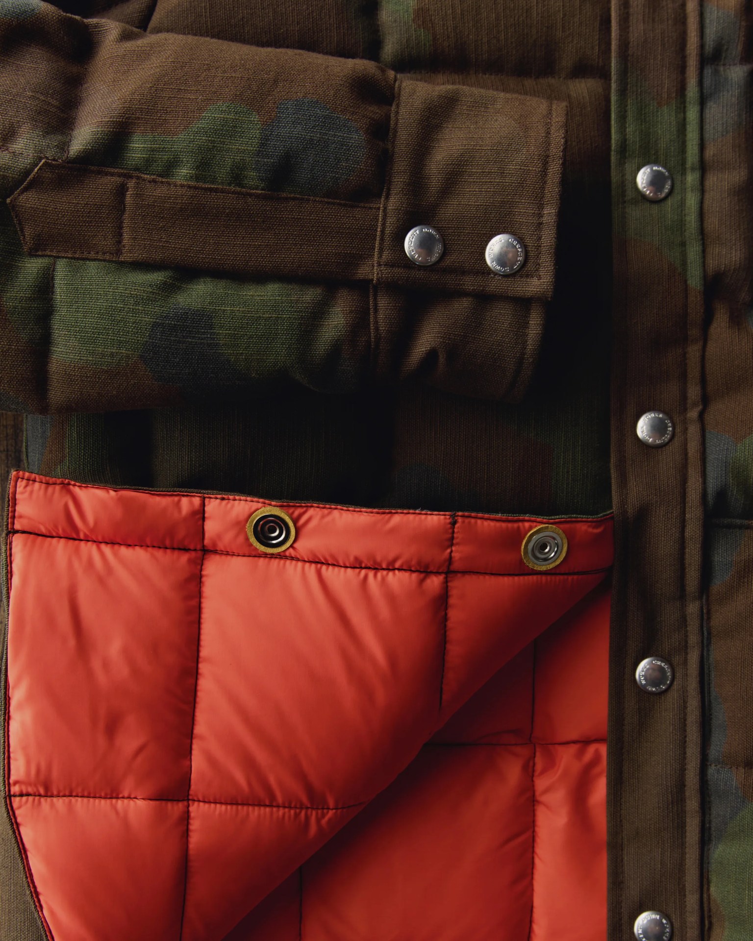 Close-up of a camouflage jacket sleeve with silver snap buttons and an orange quilted lining partially folded back.