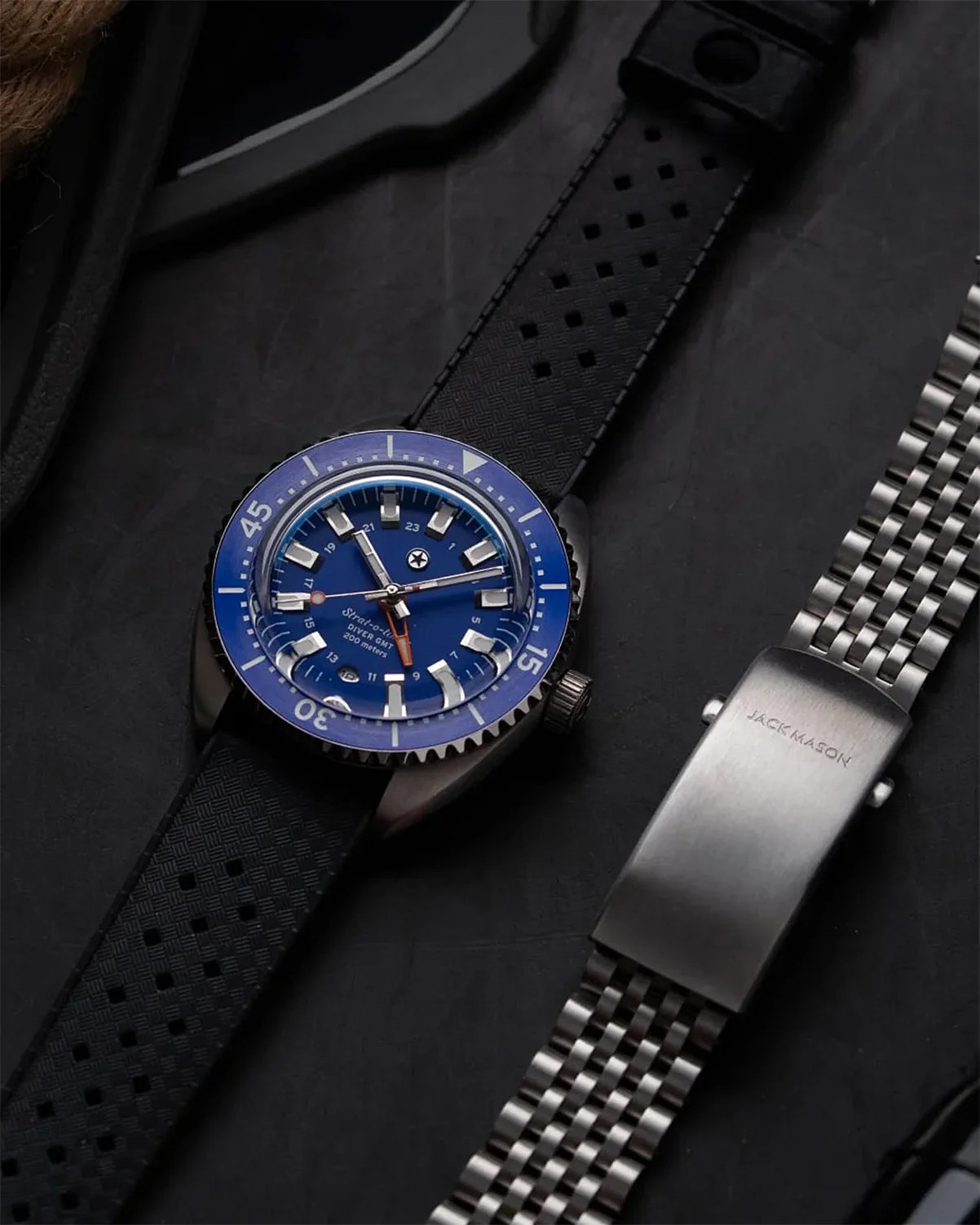 Blue diver watch with black perforated rubber strap next to a silver metal watch band on a dark surface.